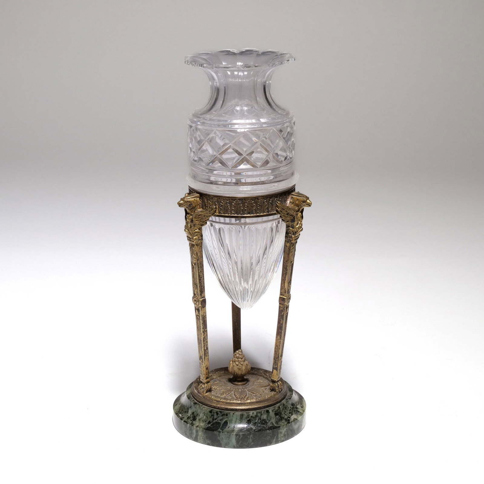 19TH CENTURY FRENCH EMPIRE STYLE CUT CRYSTAL AND CHASED BRASS VASE: The vase features a clear cut crystal body with diamond-cut patterns on the upper cylinder and fluted ribbing on the tapered lower section. It is supported by a chased brass mount with three winged ea