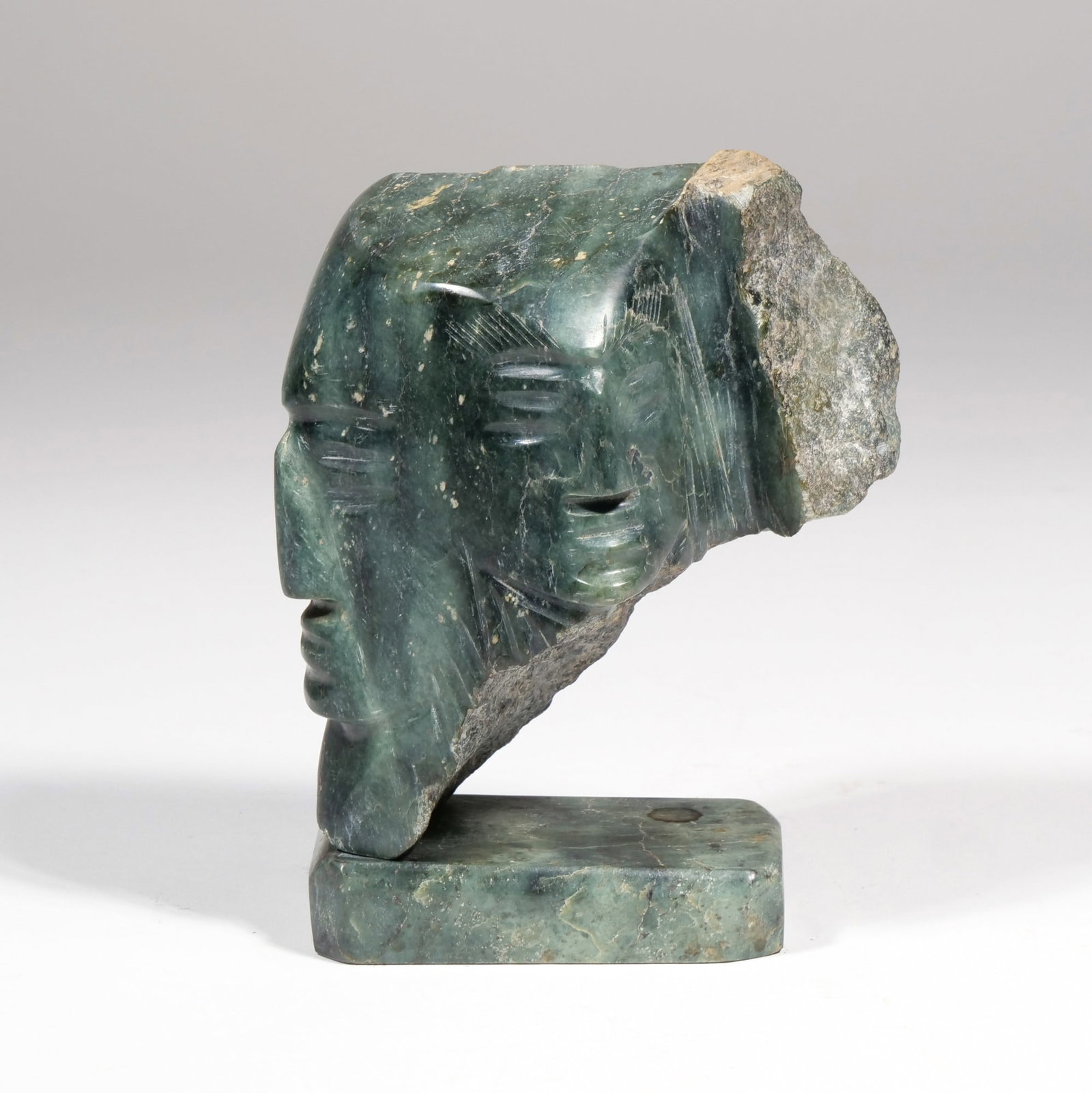 INUIT SCHOOL (20TH CENTURY) STONE CARVING: Stone Figural Head Carving Green stone h. 6.25 x w. 4.5 x d. 3.25 in. The sculpture features two stylized faces carved into dark green stone with a raw-edged reverse and is mounted on a 3.5 x 2 in.