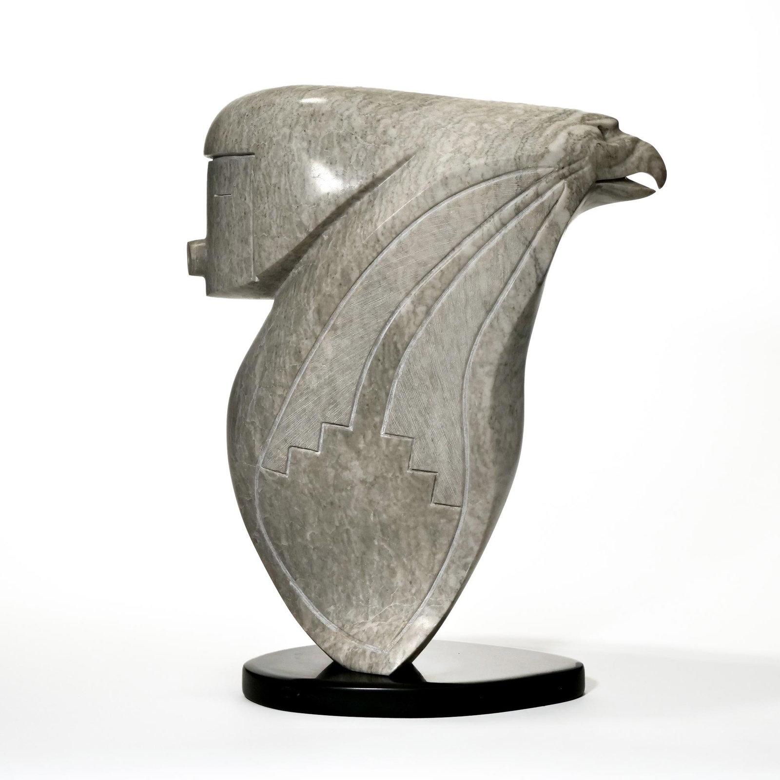 RICHARD D. TSOSIE (NAVAJO AMERICAN, 20TH/21ST CENTURY) STONE SCULPTURE: Carved from a single block of gray variegated stone and mounted on a low, oval black base. Abstracted, elongated figural form combining a rectangular, simplified human face at the front and a bird-lik