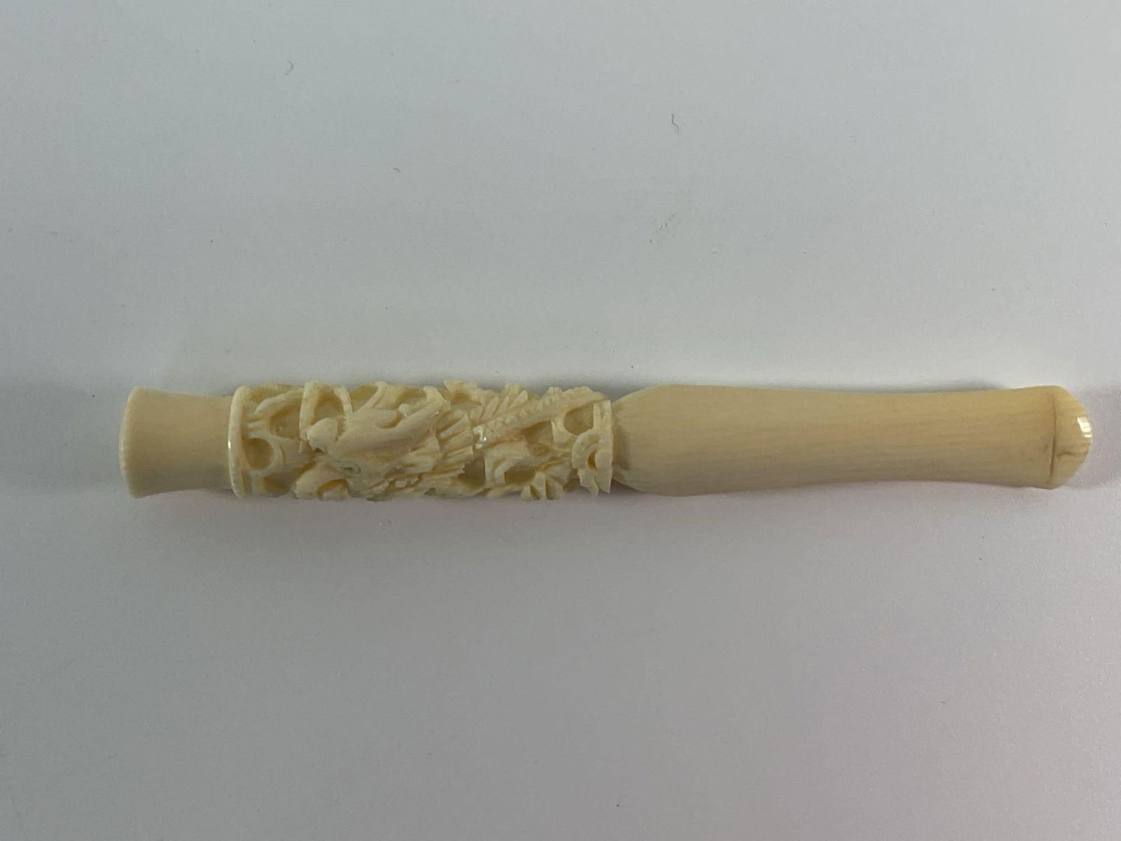 Carved Bone Cigarette Holder: A finely carved cylindrical object, possibly a cigarette holder or handle. The upper portion features intricate relief carving, possibly floral or depicting mythical creatures, showcasing detailed cra