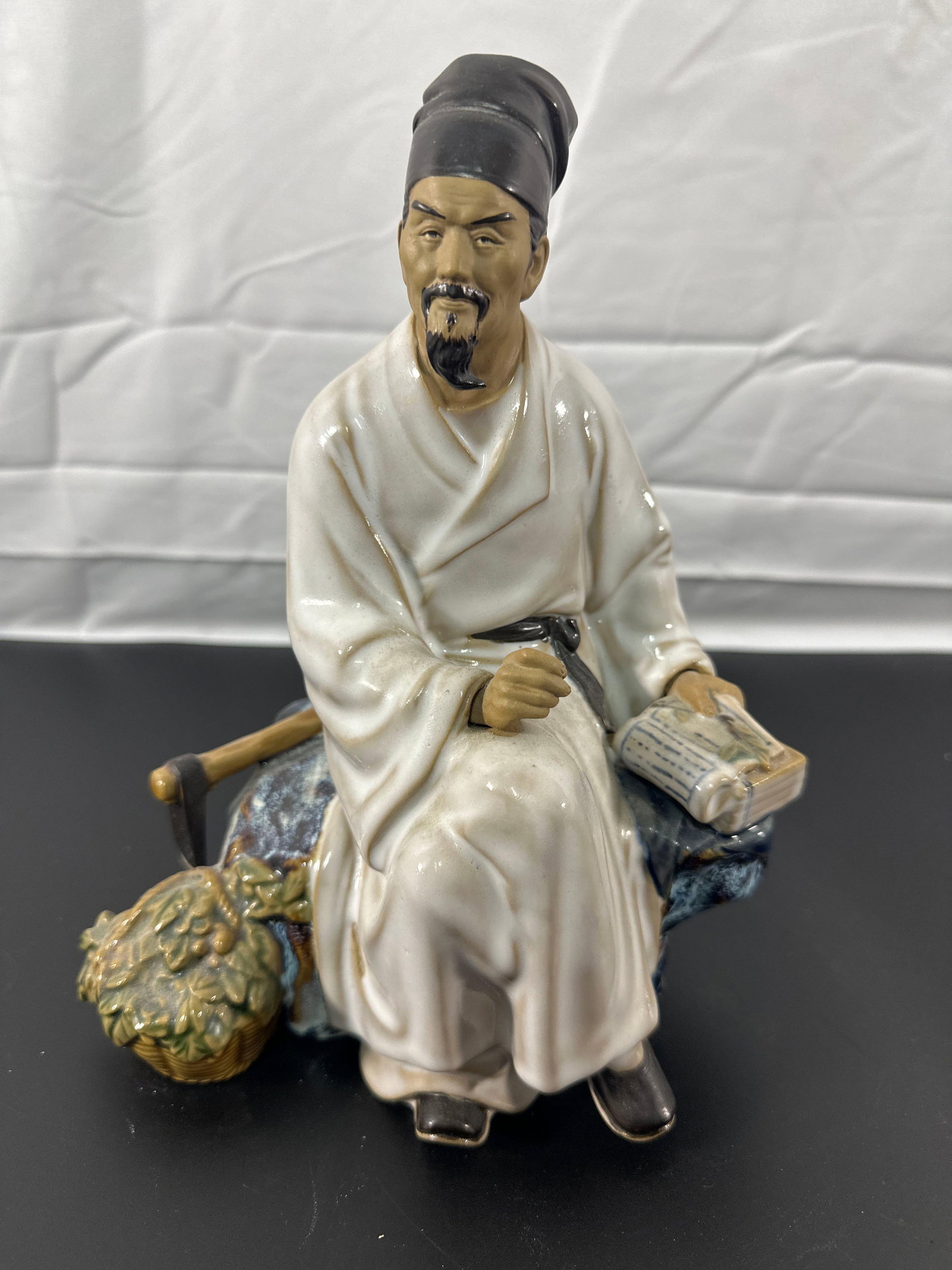 Chinese Porcelain Scholar Figurine with Scroll: This figurine represents a traditional Chinese scholar, depicted in a seated position, wearing a white robe and a classic scholar's hat. The figure holds a scroll in one hand, symbolizing knowledge, w