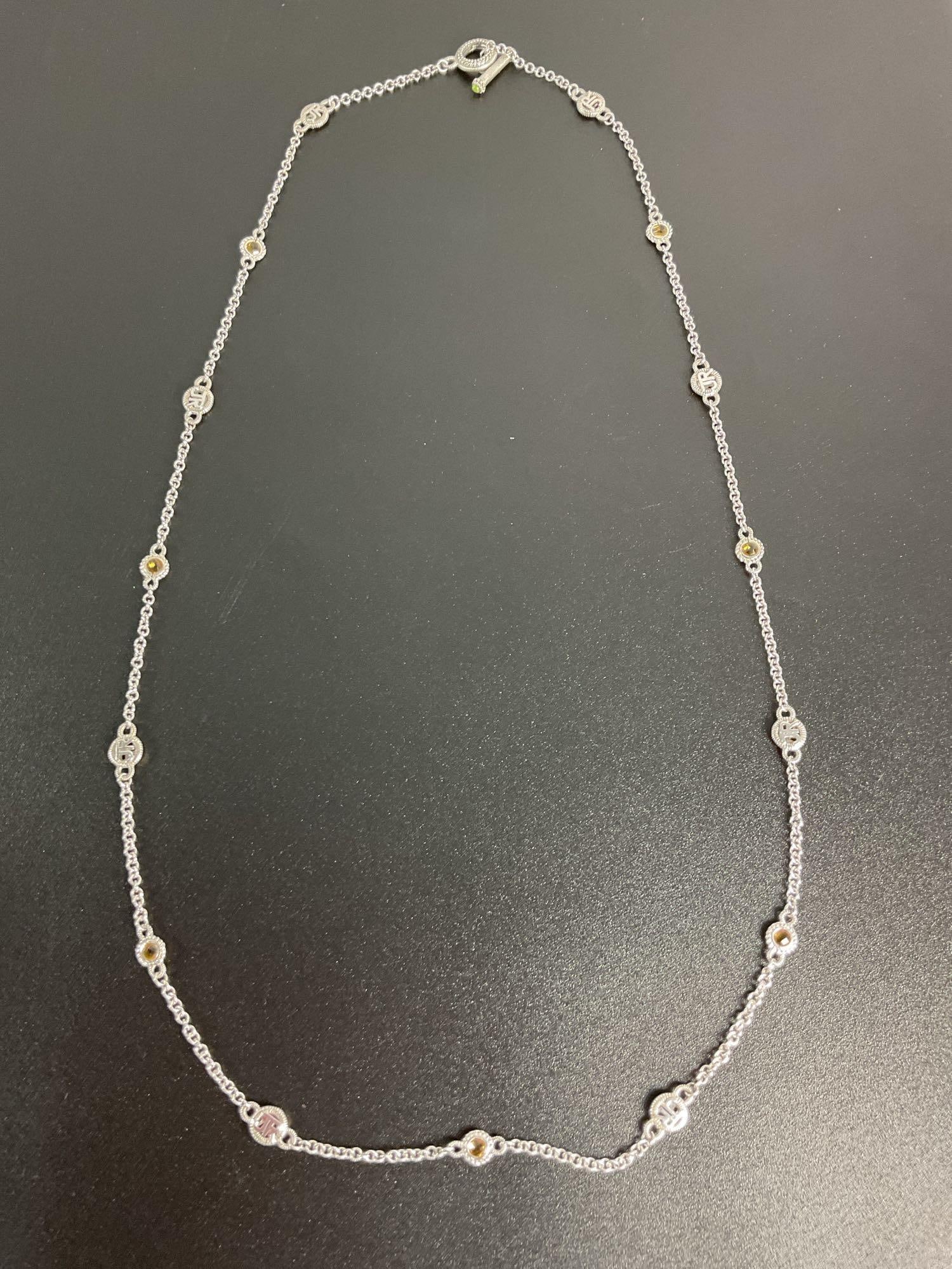 Judith Ripka Sterling Silver and Citrine Station Necklace: A beautifully crafted Judith Ripka station necklace featuring sterling silver chain links with alternating decorative medallions engraved with the "JR" logo and faceted citrine gemstone accents. The t