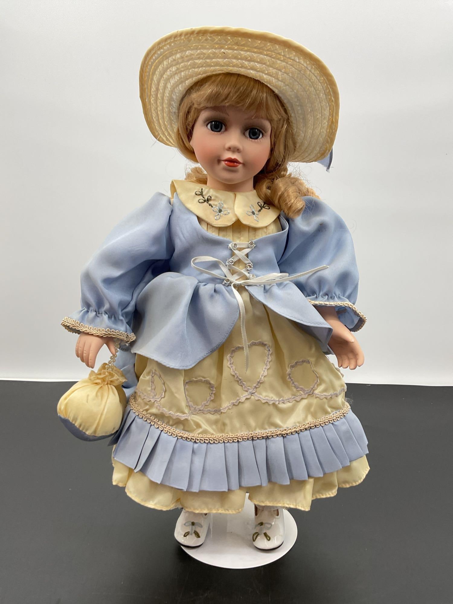 Porcelain Doll in Blue and Yellow Bonnet Dress: This porcelain doll features light brown hair styled in a side ponytail, blue eyes, and softly blushed cheeks with a gentle smile. She is dressed in a pastel blue and pale yellow gown with a pleated s