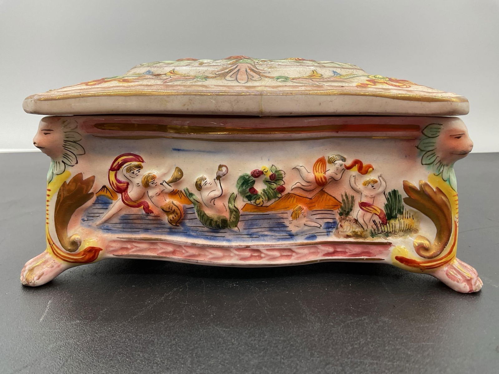 Italian Capodimonte-Style Porcelain Trinket Box: This ornate hand-painted porcelain trinket box features a raised figural scene on the lid surrounded by scrollwork and mythological cherubs or putti on all sides. The vibrant colors, baroque detailing
