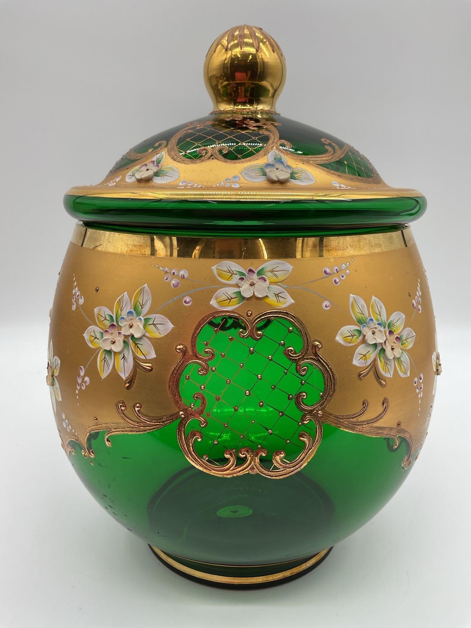 Bohemian Green Glass Lidded Jar: Handmade Czech glass jar in emerald green with gold enamel overlay, floral detailing, and applied white enamel accents. The lid is topped with a gold finial and features ornate lattice and scrollwork.