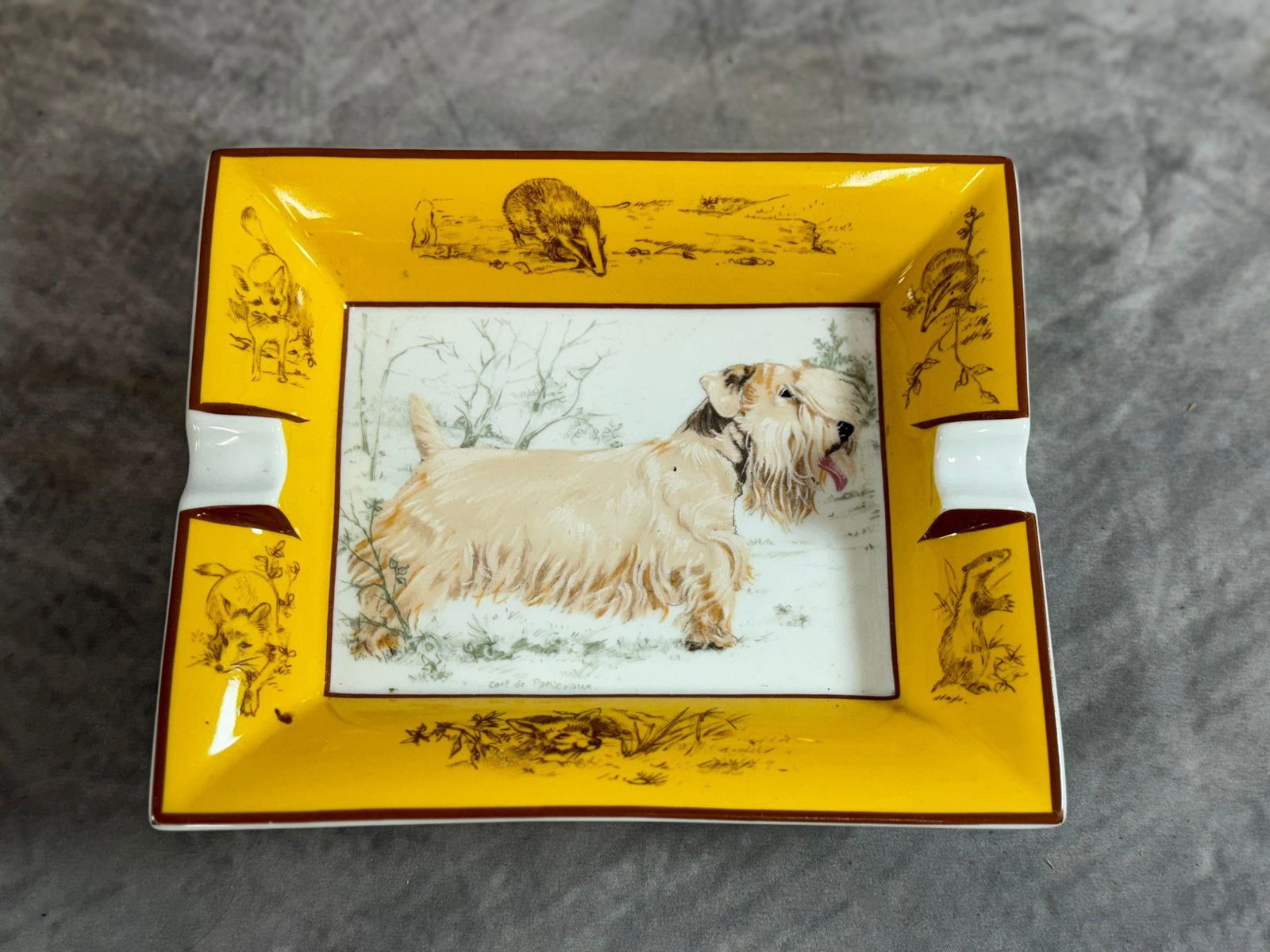 Hermes Porcelain Dog Ashtray: Elegant and collectible Hermes porcelain ashtray featuring a detailed illustration of a long-haired terrier in a naturalistic setting. The border is adorned with wildlife-themed sketches including bad