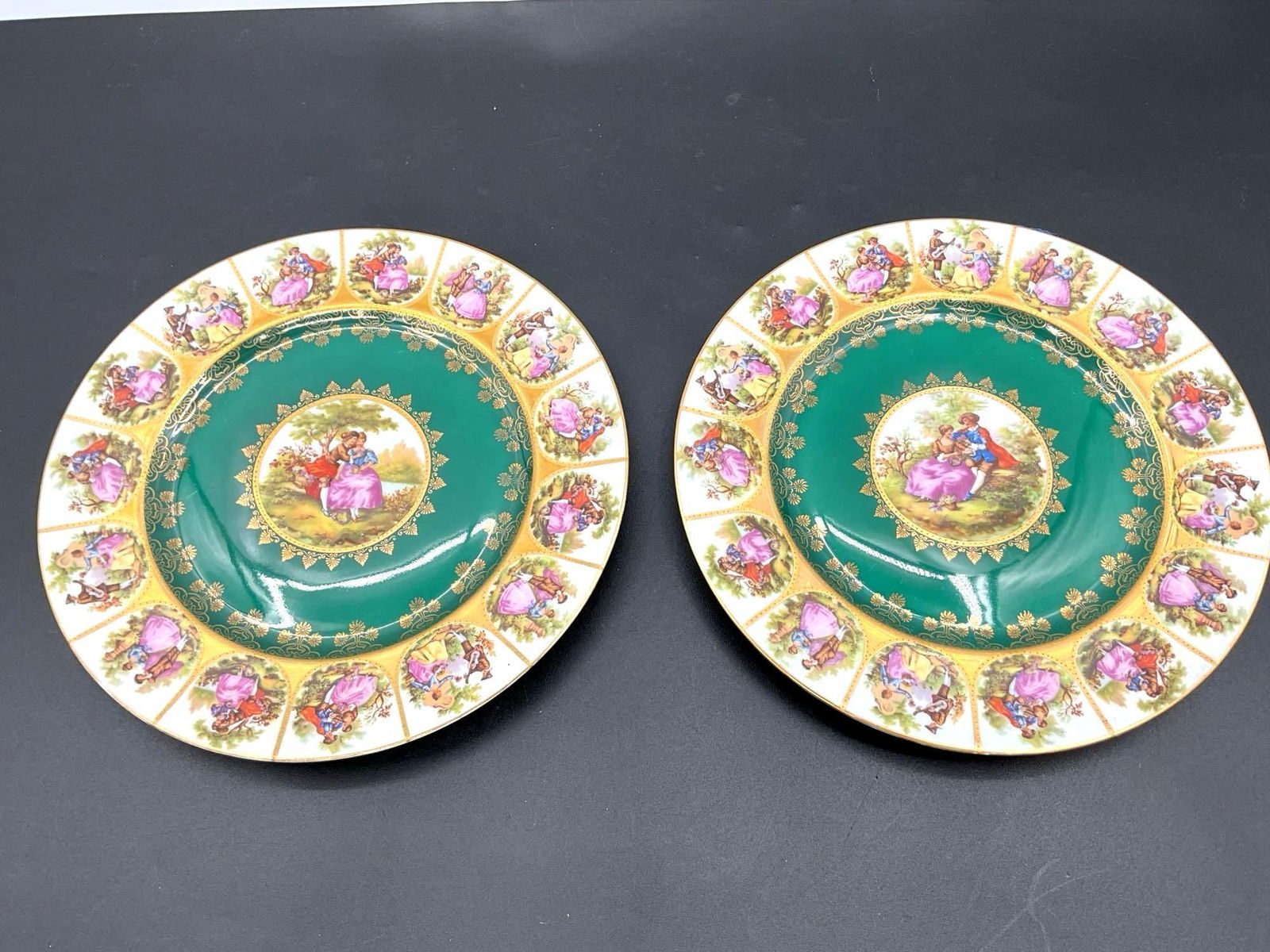 Mitterteich MM Bavaria Decorative Plates with Rococo Courting Scenes: A pair of ornate Bavarian porcelain plates featuring richly detailed Rococo-style courting scenes. The plates are decorated with a deep emerald green border, surrounded by alternating vignettes of rom