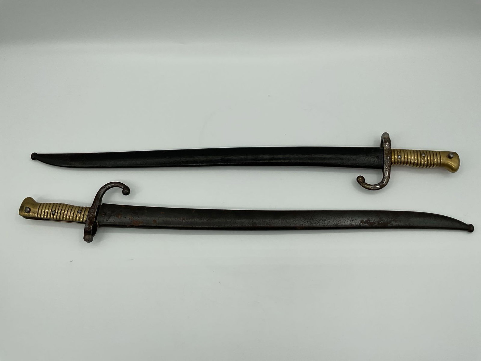 Pair of French Model 1866 Chassepot Bayonets with Scabbards: Matched pair of French Model 1866 bayonets, each featuring a full length curved blade, brass ribbed grip, steel crossguard with hooked quillon, and original metal scabbard. These yataghan style bayone