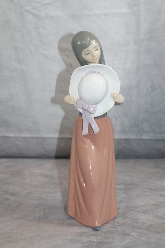 Lladro Figurine – Girl with Hat: A charming porcelain figurine by Lladro, depicting a young girl gently holding a wide-brimmed white hat adorned with a soft lavender ribbon. She wears a flowing dusty rose skirt and has long hair casc