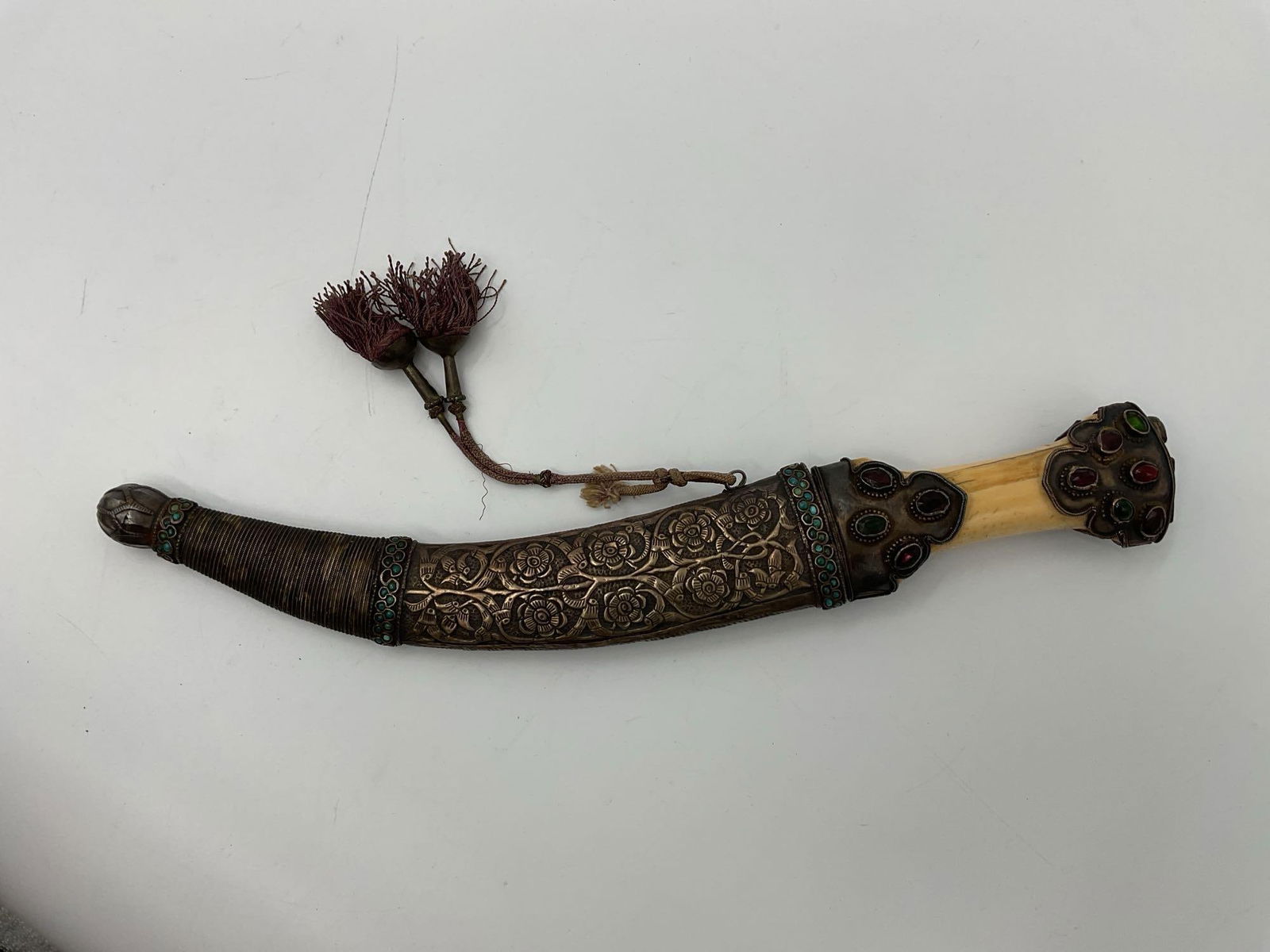 Ornate Middle Eastern or South Asian Khanjar Dagger: Curved ceremonial dagger featuring a highly decorated sheath and hilt, adorned with colorful glass or semi-precious cabochon stones, floral embossing, and silver-tone metalwork. The handle appears to