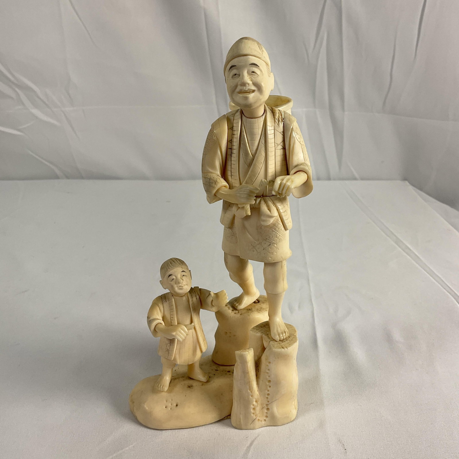 Carved Japanese father and child figurine: Intricately detailed sculpture of a smiling man in traditional Japanese attire carrying a basket on his back, accompanied by a young child holding his hand. Both figures are barefoot and depicted mid-