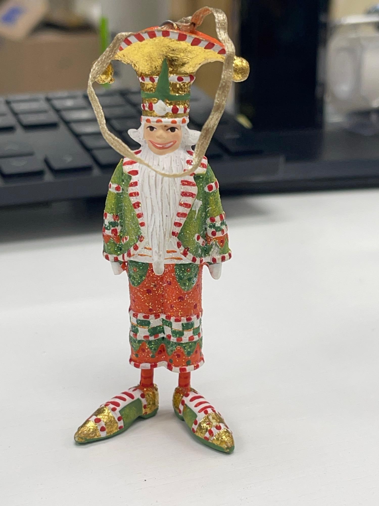 Nutcracker Mini Ornament by Patience Brewster for MacKenzie-Childs: Festive hand-painted mini resin ornament of a nutcracker in a whimsical red, green, and gold outfit, complete with an exaggerated hat and curled shoes. A charming holiday collectible by Patience Brews