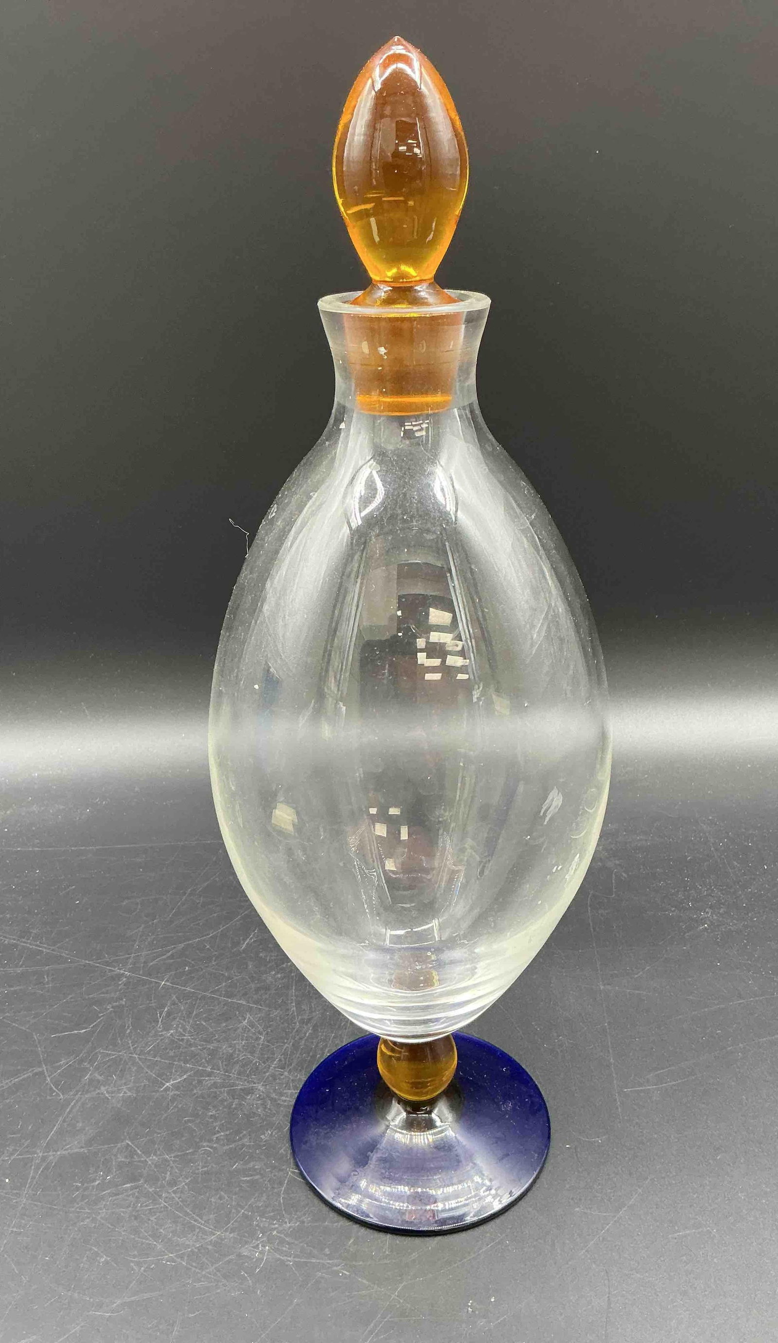 Murano Style Art Glass Apothecary Jar with Amber Finial: Description: Tall blown art glass jar featuring an elegant clear oval body, amber finial and base accent, and cobalt blue foot. Likely Murano style, with modern decorative flair. Medium: Blown art gla