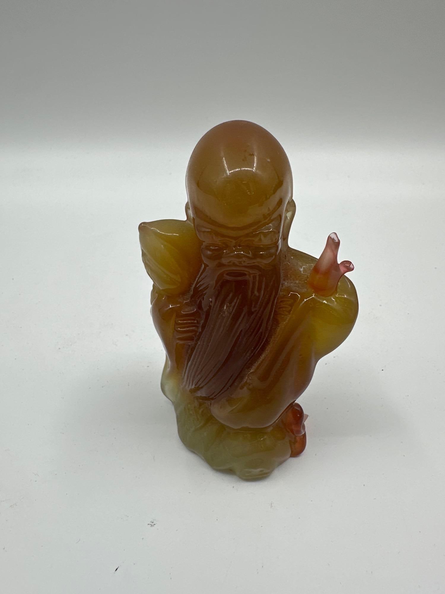 Carved Agate-Style Chinese Scholar Figurine: A small figure of a Chinese sage or immortal, carved in translucent agate-like stone with warm honey, amber, and green hues. The figure holds a staff and peach, symbolizing longevity. Medium: Polished