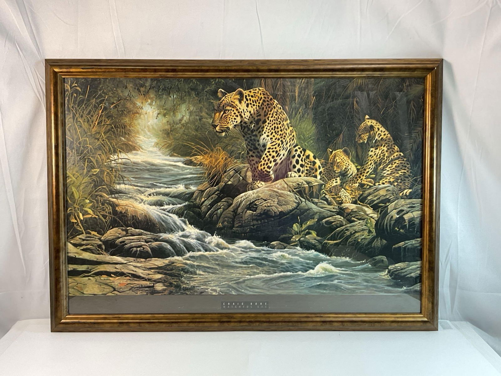 Craig Bone (American, b.1955) Lithograph: The piece is titled "Watchful Eye".This is a framed wildlife print featuring a leopard family near a flowing river, set in a dense jungle or forest. The composition is highly detailed, with re