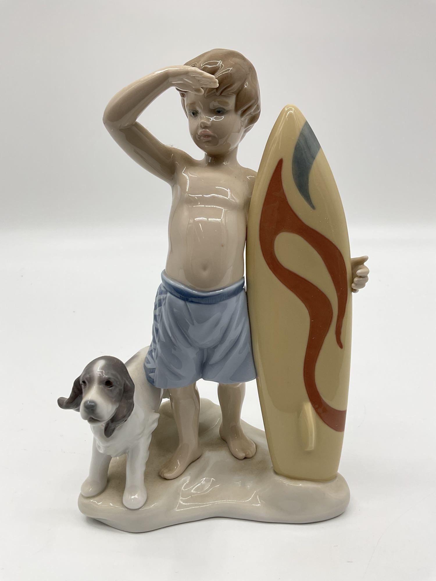 Lladro Figurine – Surfs Up 01008110: Playful porcelain figurine by Lladro depicting a young boy in blue swim trunks shielding his eyes while standing beside a yellow surfboard with red detailing. A loyal dog stands next to him, completin