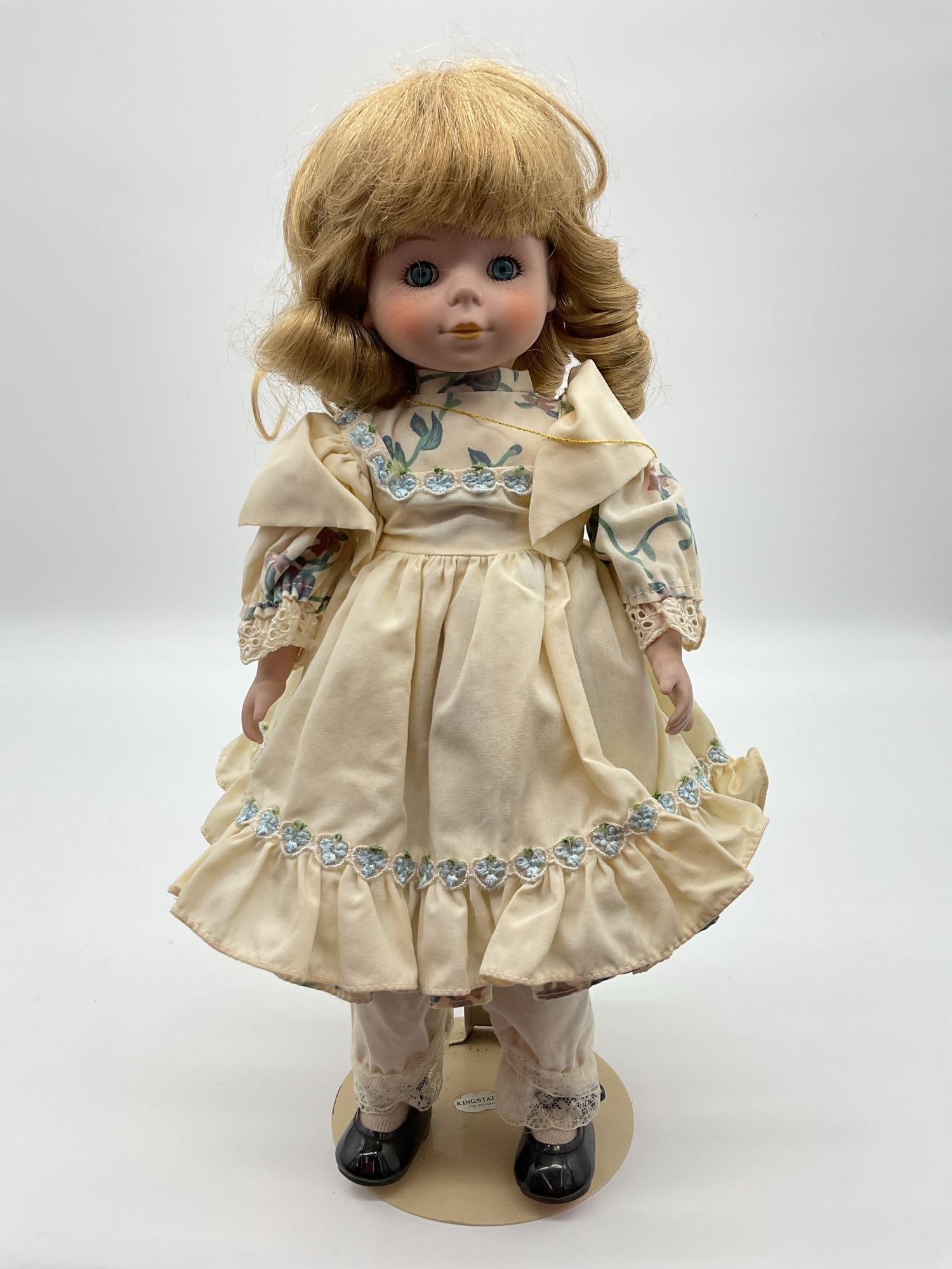 Vintage Kingstate Porcelain Doll: This vintage porcelain doll is dressed in an antique-style cream-colored dress with blue floral embroidery and lace accents. She has blonde hair styled in soft waves. Her hands are covered with lace g