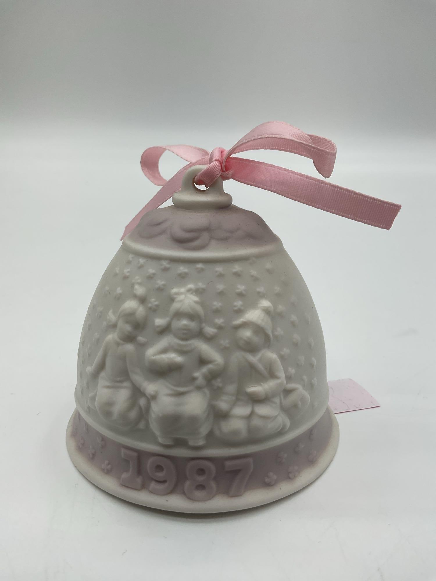 Lladro 1987 Christmas Bell Ornament: A matte porcelain bell featuring three caroling children in relief, topped with a pink ribbon, and dated 1987. Dimensions: H3in Dia3in Medium: Unglazed (matte) porcelain