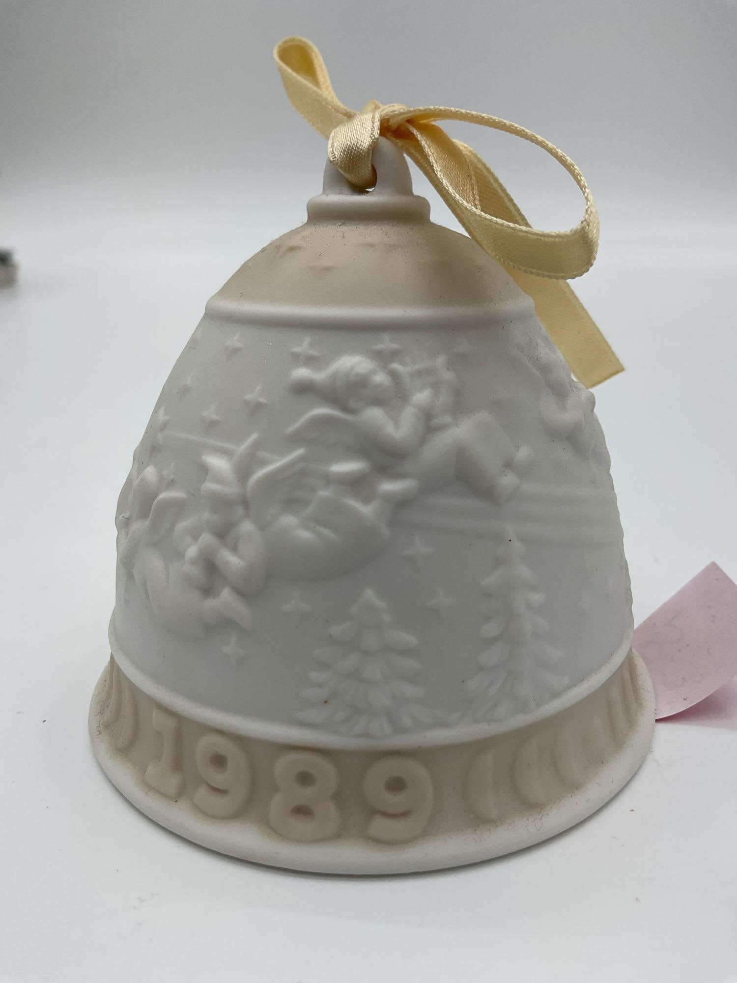 Lladro 1989 Christmas Bell Ornament: A matte porcelain bell featuring Santa in his sleigh flying over snowy pine trees, topped with a gold ribbon, and dated 1989. Dimensions: H3in Dia3in Medium: Unglazed (matte) porcelain