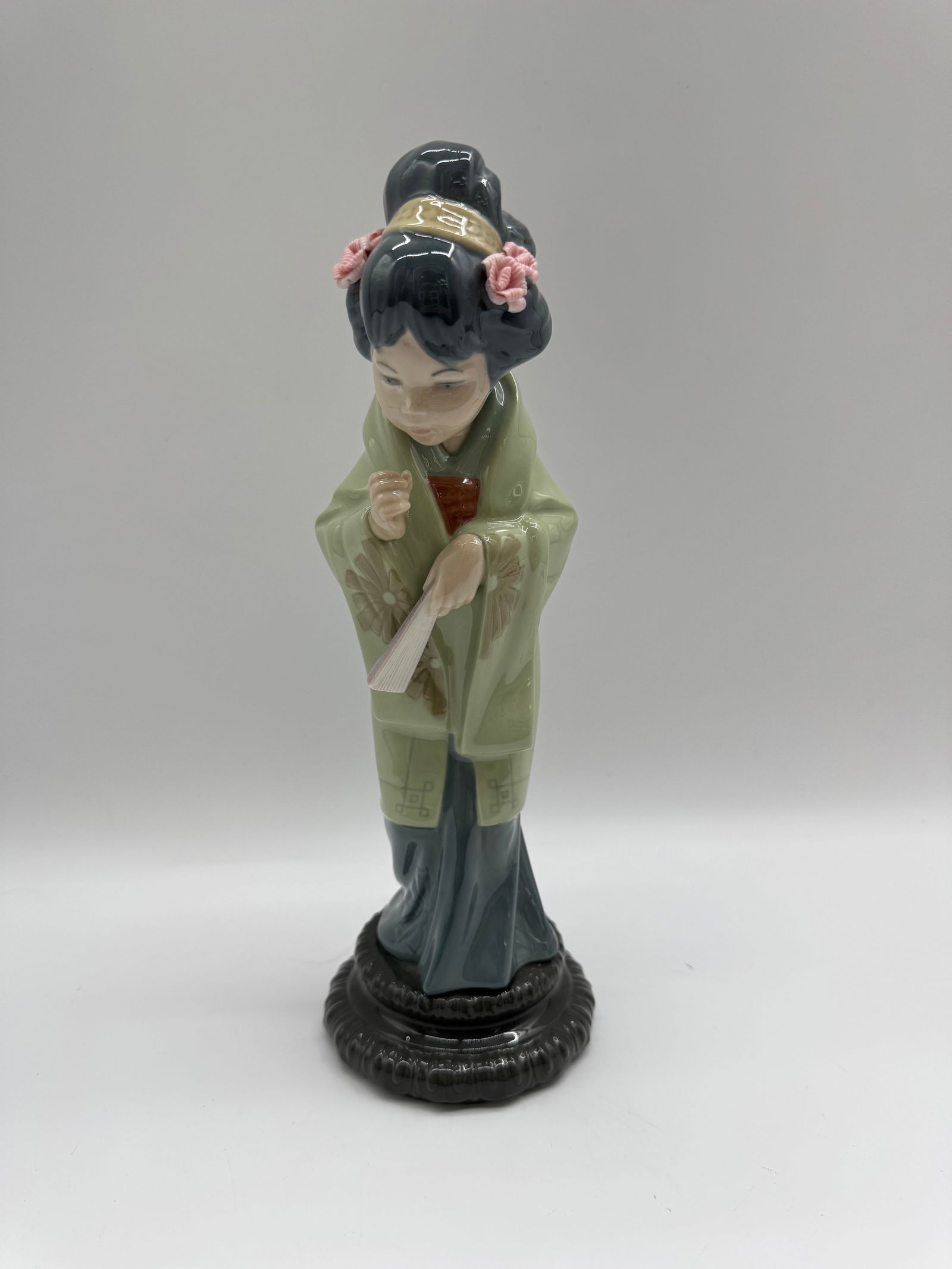 Vintage Japanese Geisha Lladro: Vintage Japanese figurine depicting a geisha girl in traditional dress. The piece features detailed ceramic work and stands on a decorative base. The geisha is holding a fan and adorned with pink flow
