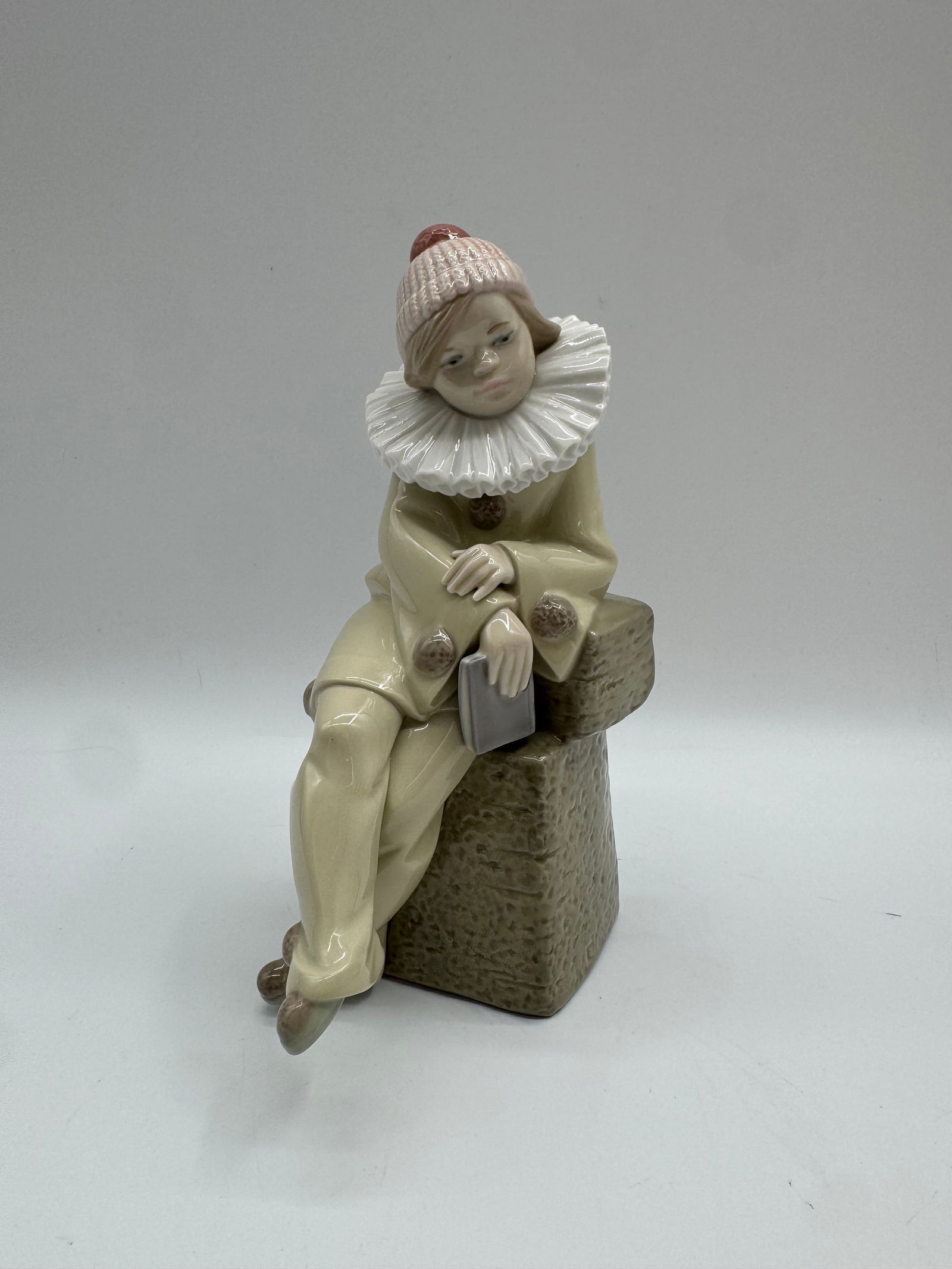 Lladro Porcelain Figurine - Resting Clown with Book: Lladro porcelain figurine of a clown resting on a block, holding a book. Marked on the base. Measures approximately 6 inches tall.