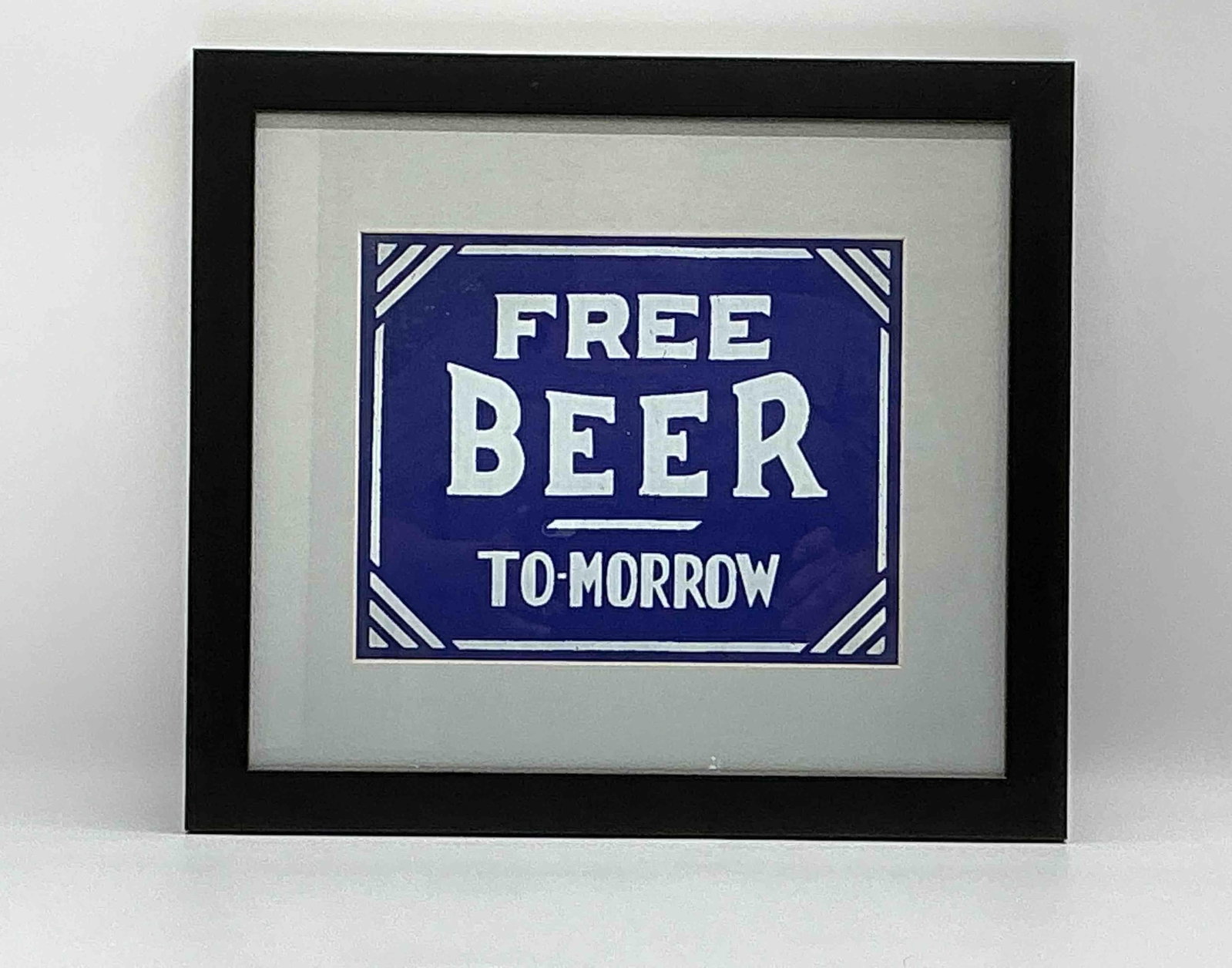 Framed Sign Print "Free Beer To-morrow": Humorous novelty print reading Free Beer To-morrow in bold white lettering against a blue background with decorative border. Framed under glass with a simple black frame. Popular as vintage-