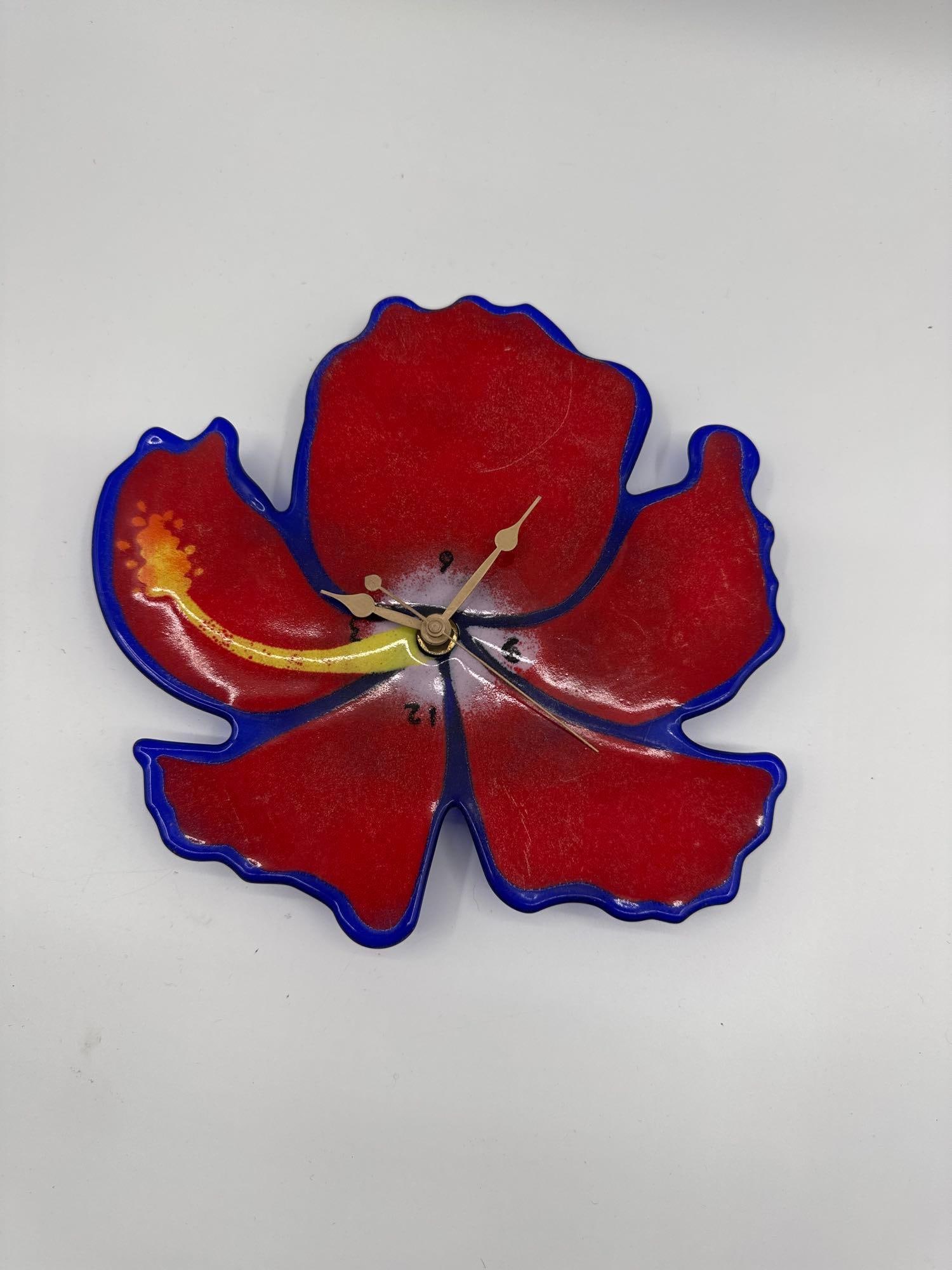Red Flower Wall Clock: Whimsical wall clock shaped like a red flower with blue edges and yellow detail, featuring minimalist time markings.Medium: Glazed ceramic with metal clock hands and quartz movement