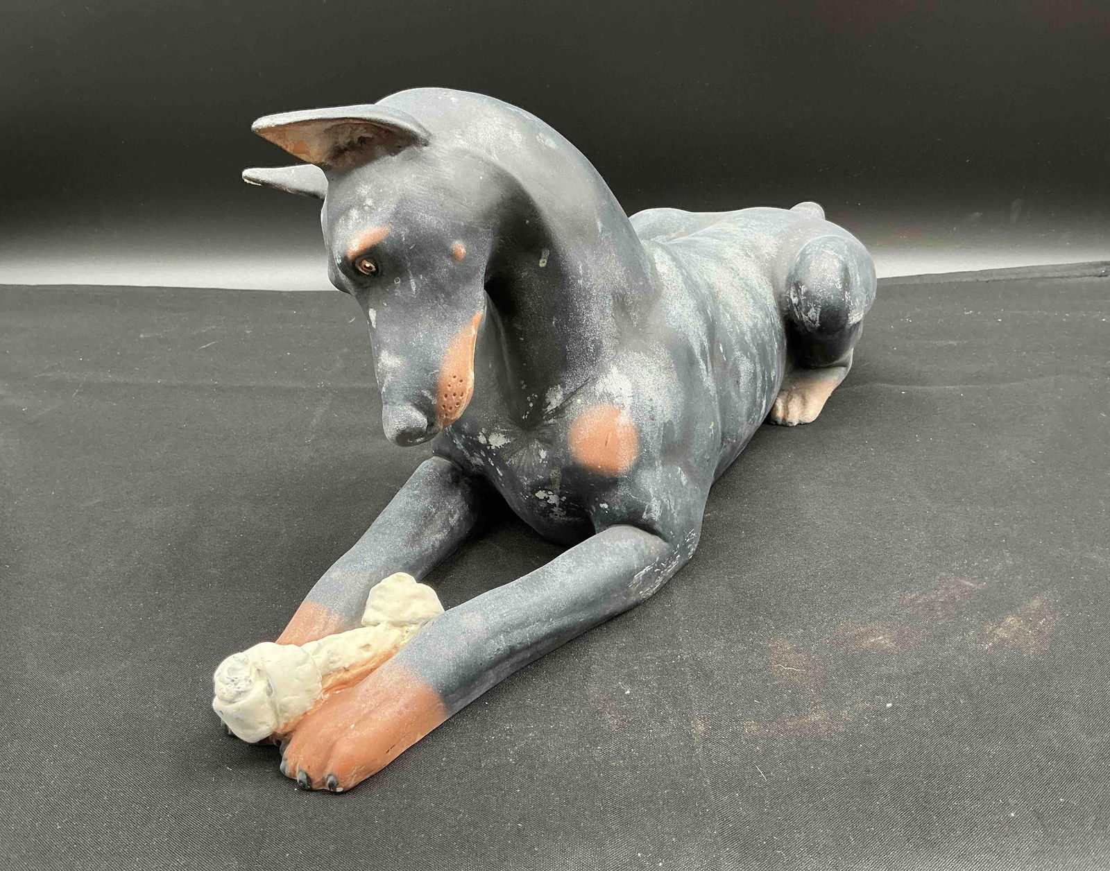 Whimsical Doberman Figurine Holding Bone: Hand-painted ceramic figurine of a stylized Doberman Pinscher lying down and gently holding a white bone in its paw. Matte finish with black, tan, and white accents. Medium: Painted ceramicSome wear v