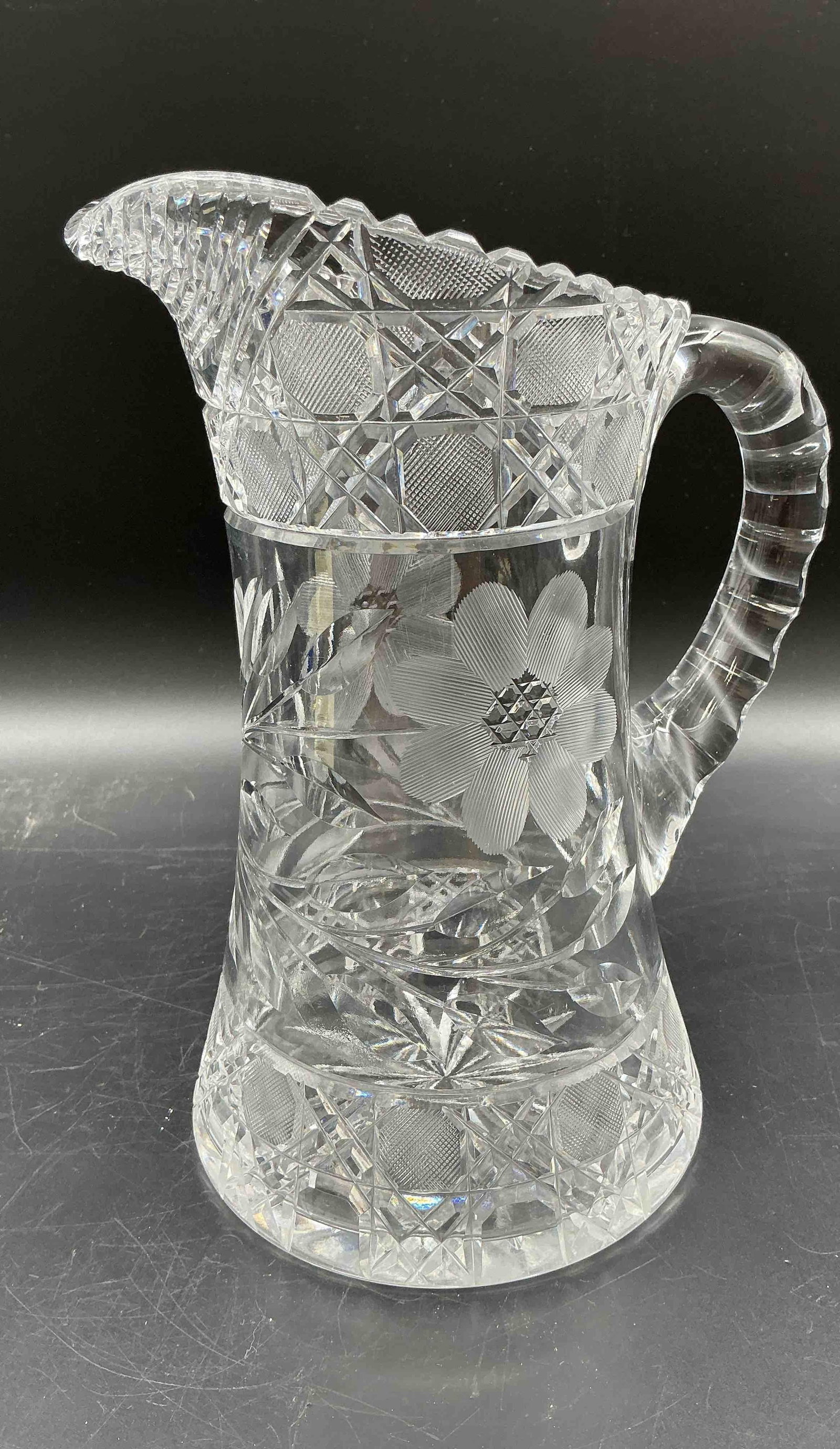 American Brilliant Style Cut Glass Pitcher with Floral Motif: Large clear glass pitcher with deep geometric cut patterns and a central etched floral motif. Features a heavily faceted handle and scalloped rim. In the style of American Brilliant Period cut glass, 