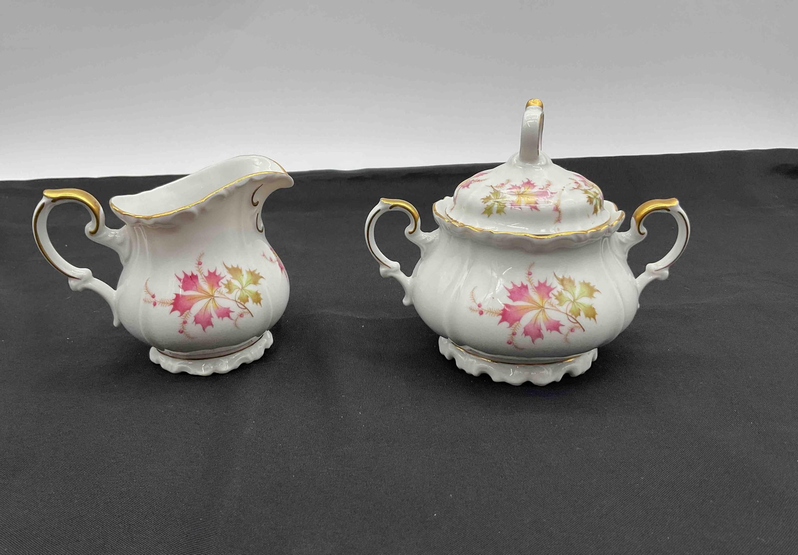 Edelstein Porcelain Creamer and Sugar Bowl Set : White porcelain creamer and lidded sugar bowl decorated with pink and green leaves and gold accents Medium: porcelain with gold detailing Dimensions: creamer H4in L5in W3in, sugar H5in L6in W4in