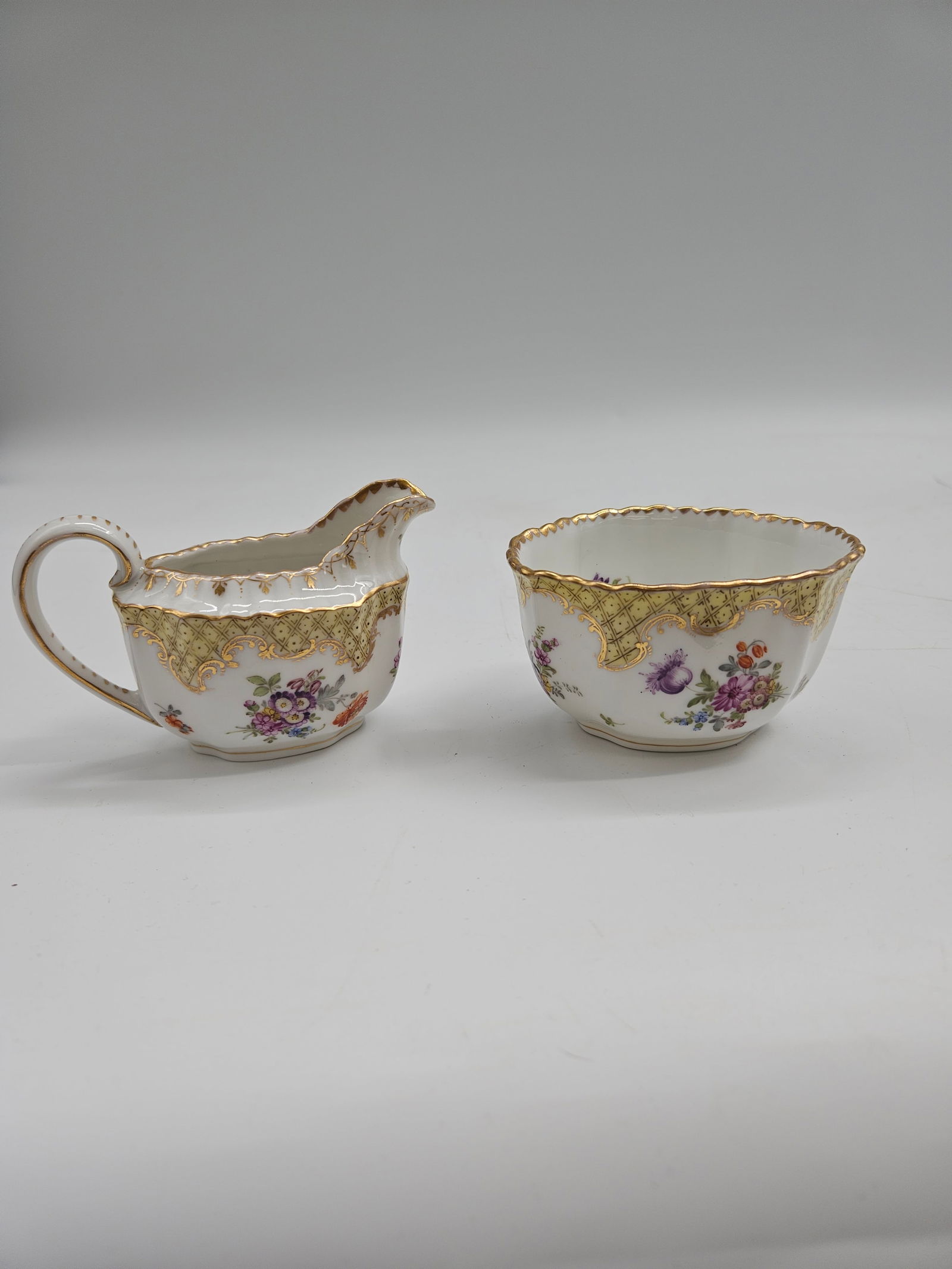 Staffordshire Floral Creamer and Sugar Bowl Set: Elegant Staffordshire porcelain creamer and sugar bowl set adorned with hand-painted purple and pink floral designs. Each piece is accented with intricate gold gilt borders and scalloped rims, reflect