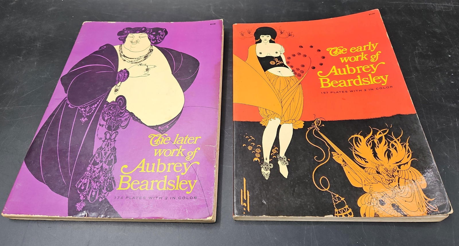 The Early and later Works of Aubrey Beardsley Book Lot: Two vintage paperback books showcasing the art of Aubrey Beardsley. The covers feature bold illustrative designs in purple and orange tones. Both volumes highlight the distinctive and influential styl
