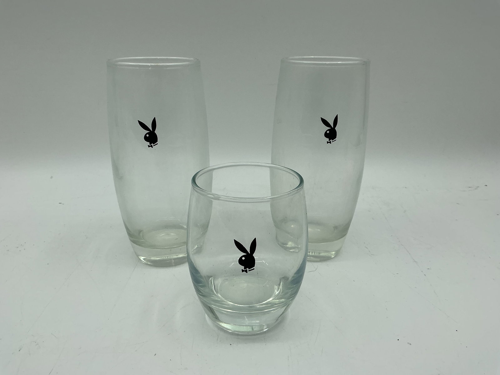 Playboy Bunny Logo Glassware Set (3): Set of three clear glass tumblers featuring the Playboy bunny logo in black. Includes two taller glasses (6in x 3in x 2.5in) and one smaller glass (3.5in x 3in x 2.5in).