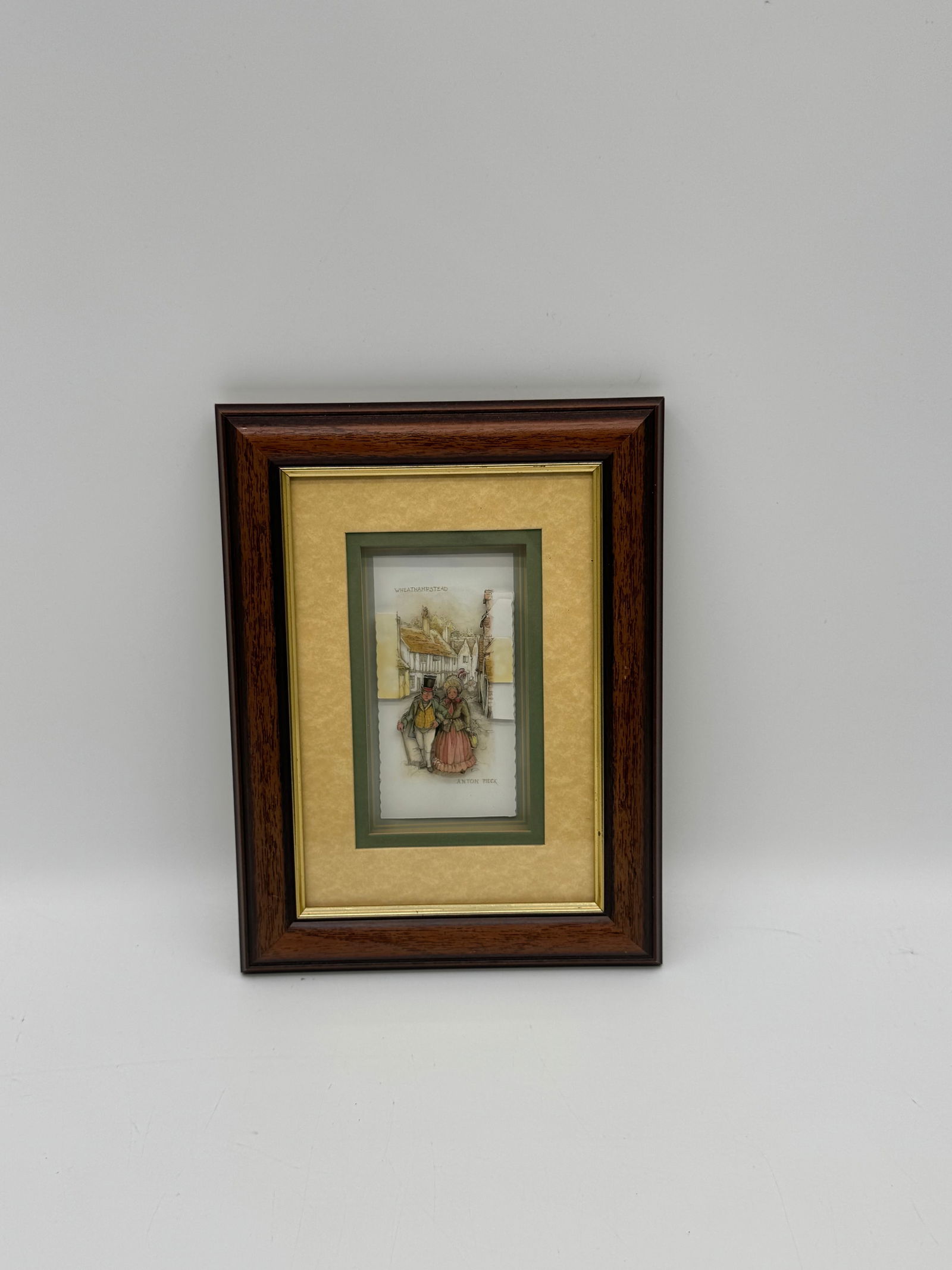 Framed Shadow Box by Anton Pieke: Framed 3D shadow box artwork by Anton Pieck depicting a charming street scene with a couple strolling past buildings. Features a dark wood frame with gold and green matting. Approx. 10 x 7.5 inches.