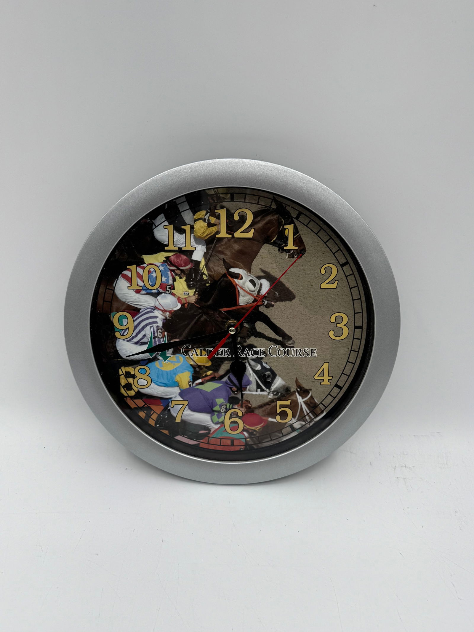 Calder Horse Racing Wall Clock - Miami Gardens: Round wall clock featuring a horse racing scene on the face. The clock has a silver-tone frame and measures approximately 11 inches in diameter. Battery operated.