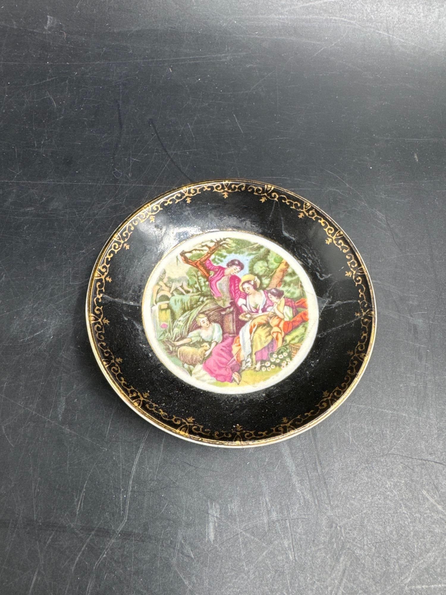 Vintage Decorative Porcelain Plate with Romantic Scene:  Small round porcelain plate featuring a central hand-colored transfer of a romantic 18th-century style courting scene, framed by a black border with intricate gilt scrollwork. A collectible deco