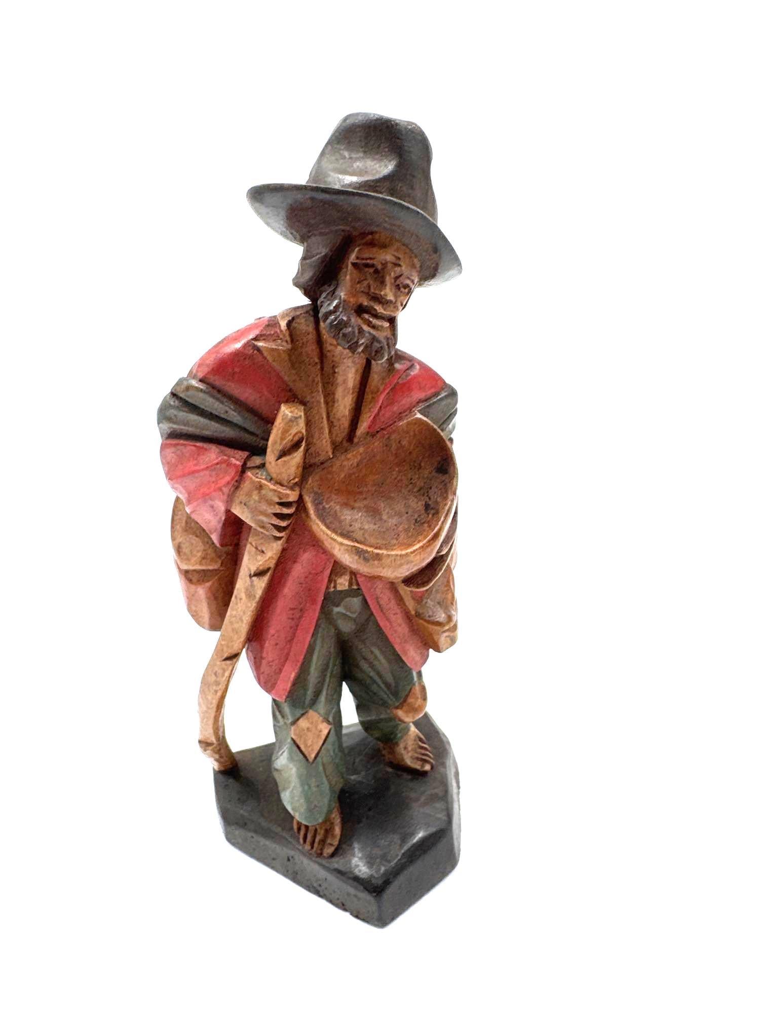Hand-Carved Figure of a Beggar with Hat and Bowl:  Painted wood sculpture of a barefoot beggar holding a staff and a bowl, wearing a patched cloak and wide-brimmed hat. Finely detailed with expressive facial features. Medium: Hand-carved and han