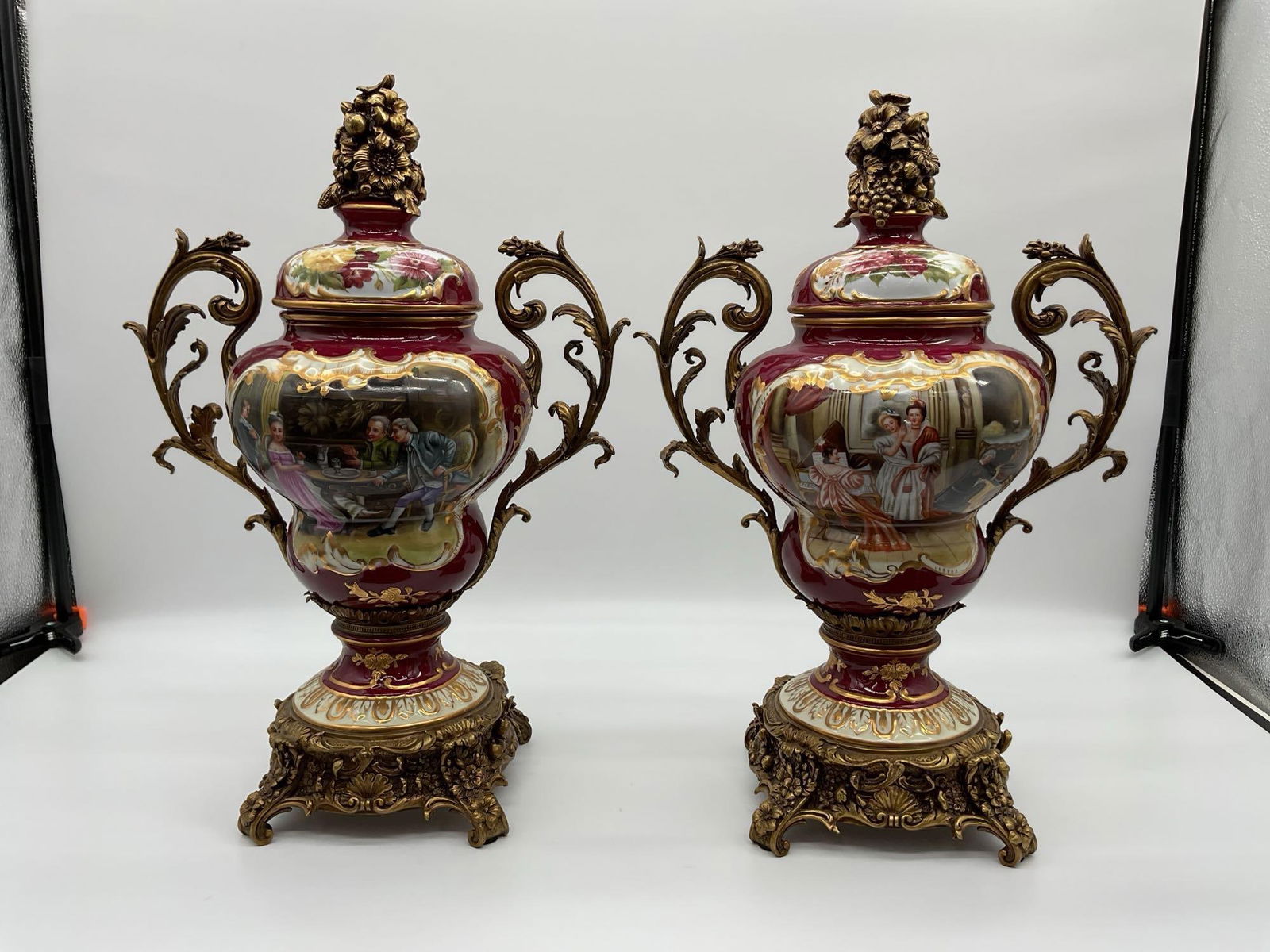 Pair of French Style Porcelain and Gilt Bronze Urns: Ornate matching covered urns with deep burgundy glaze and hand-painted romantic scenes. Adorned with floral and figural bronze finials, scrolling gilt bronze handles, and decorative bases in Rococo st