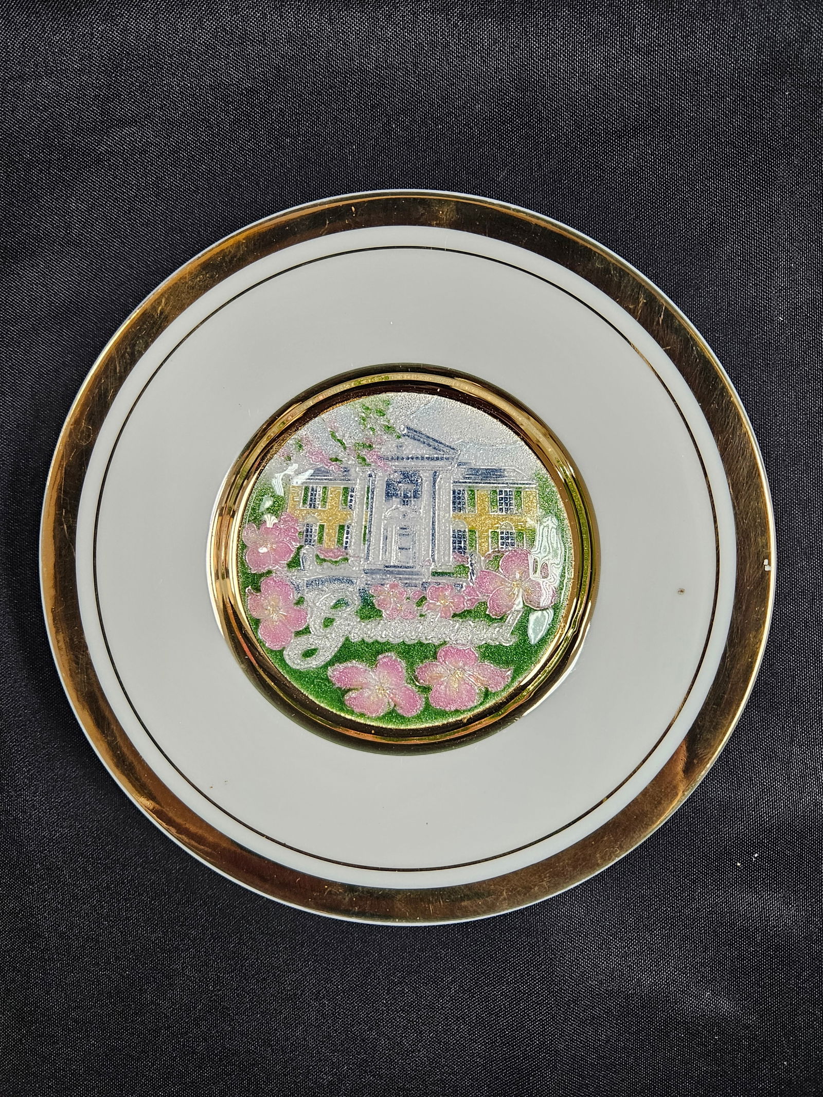Japanese Cloisonne Plate - Graceland Design: Vintage Japanese Cloisonne plate with a 24K gold edge. The plate features a central design of Graceland surrounded by pink blossoms on a green background. The back of the plate has a sticker describin