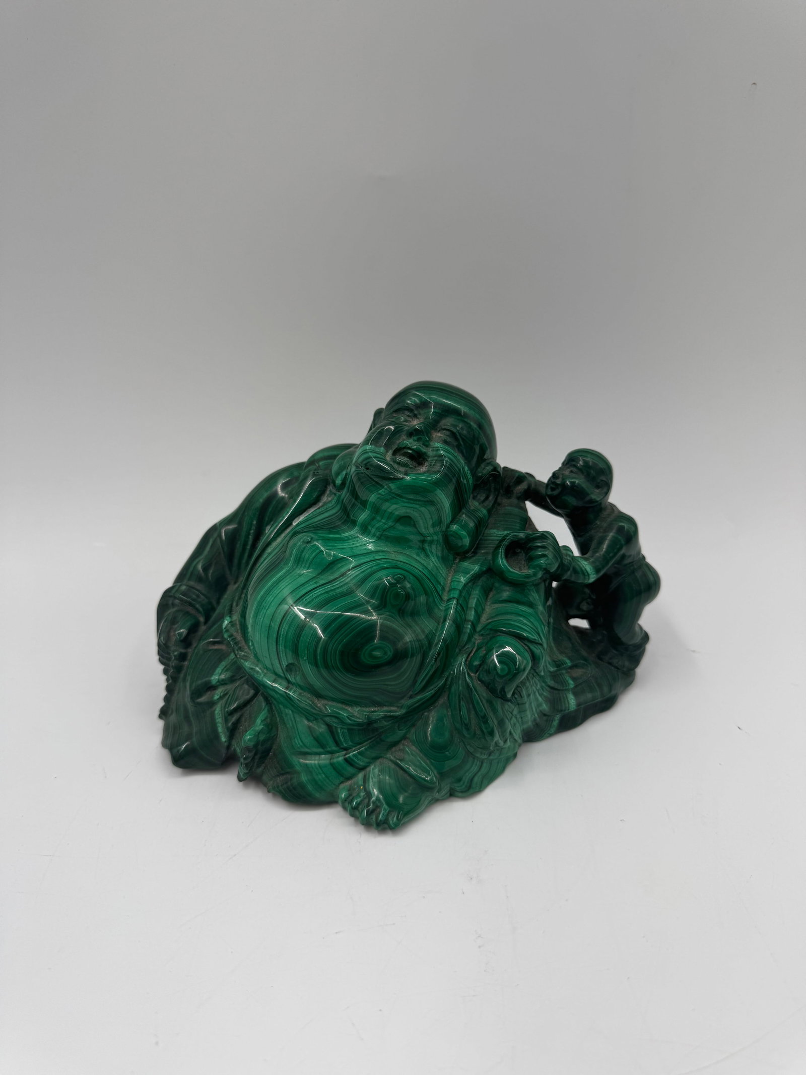 Malachite Buddha Sculpture with Child: Carved malachite sculpture depicting a seated Buddha figure with a child. The piece features intricate details and natural banding patterns in the stone. Dimensions: 7 x 4.5 inches.