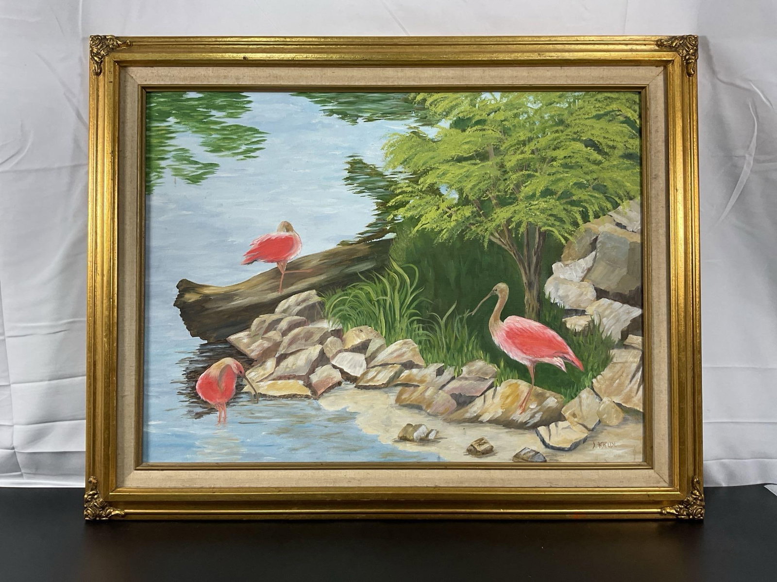 Serenity by the Water by J. Kruse: This charming painting depicts a tranquil waterside scene with vibrant pink flamingos (or possibly roseate spoonbills) standing gracefully among rocks and lush greenery. The artist has captured the ca
