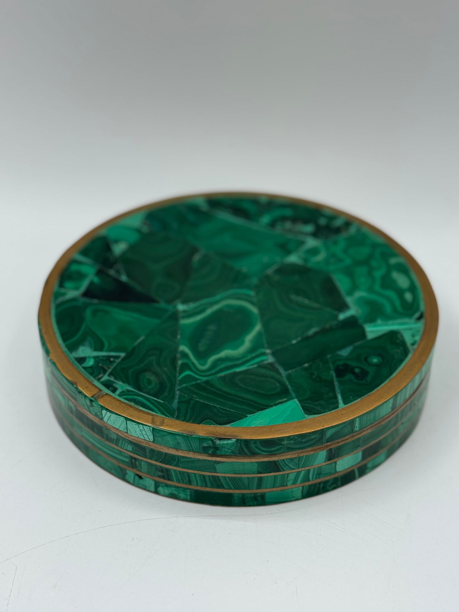 Malachite Inlaid Round Box with Brass Trim: Round box featuring malachite inlay on the lid and sides, accented with brass trim. The interior is lined with green felt. Measures approximately 2 inches tall and 8 inches in diameter. A decorative p