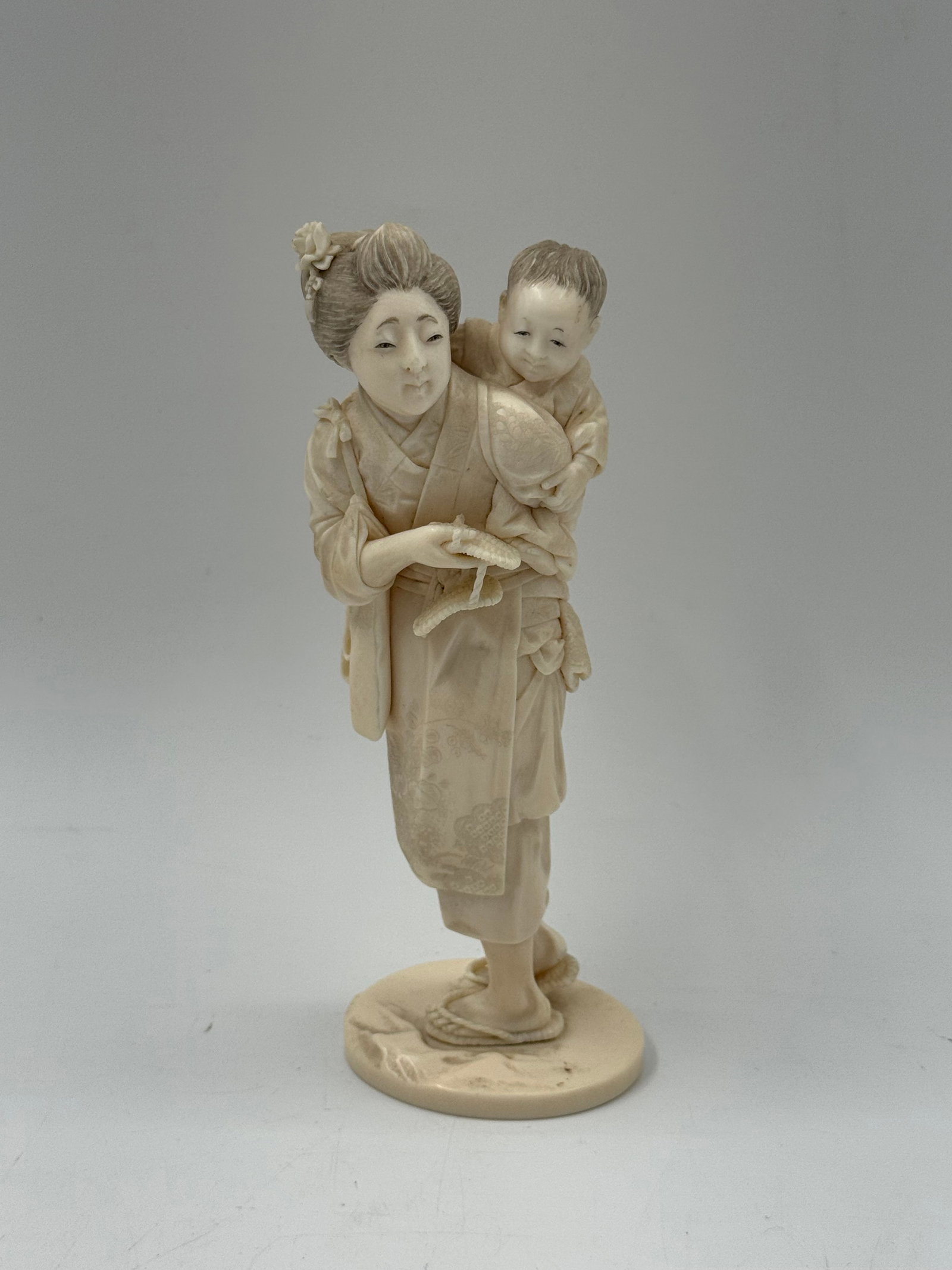 Japanese Carved Bone Okimono of Mother and Child: A carved bone okimono depicting a standing woman with a child on her back. The figure is mounted on a circular base. Measures approximately 7 x 3 inches.