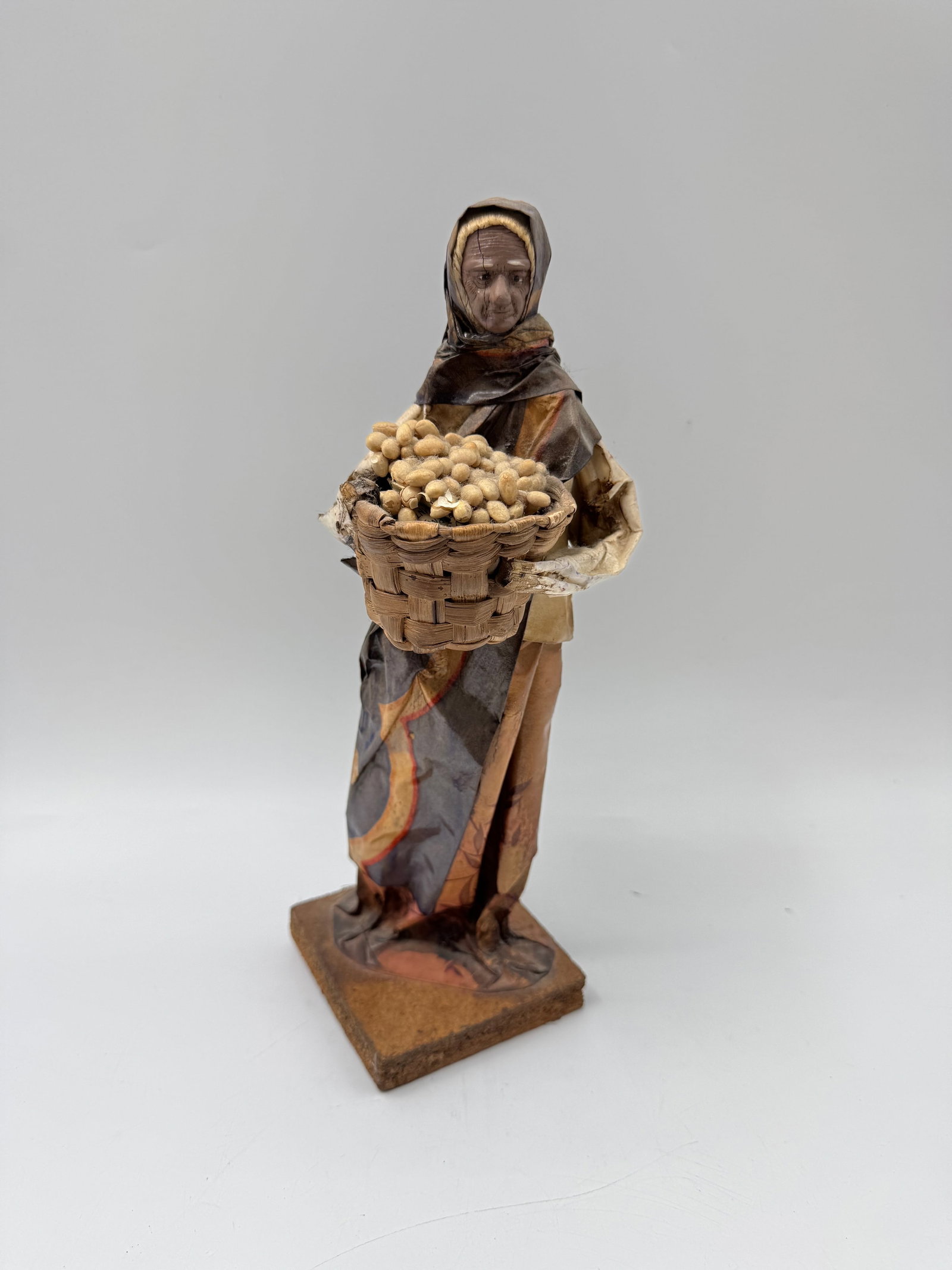 Folk Art Paper Mache Woman with Basket: Paper mache figure of a woman holding a basket filled with goods. The figure is mounted on a wooden base. Dimensions: 12 x 4 x 4 inches.