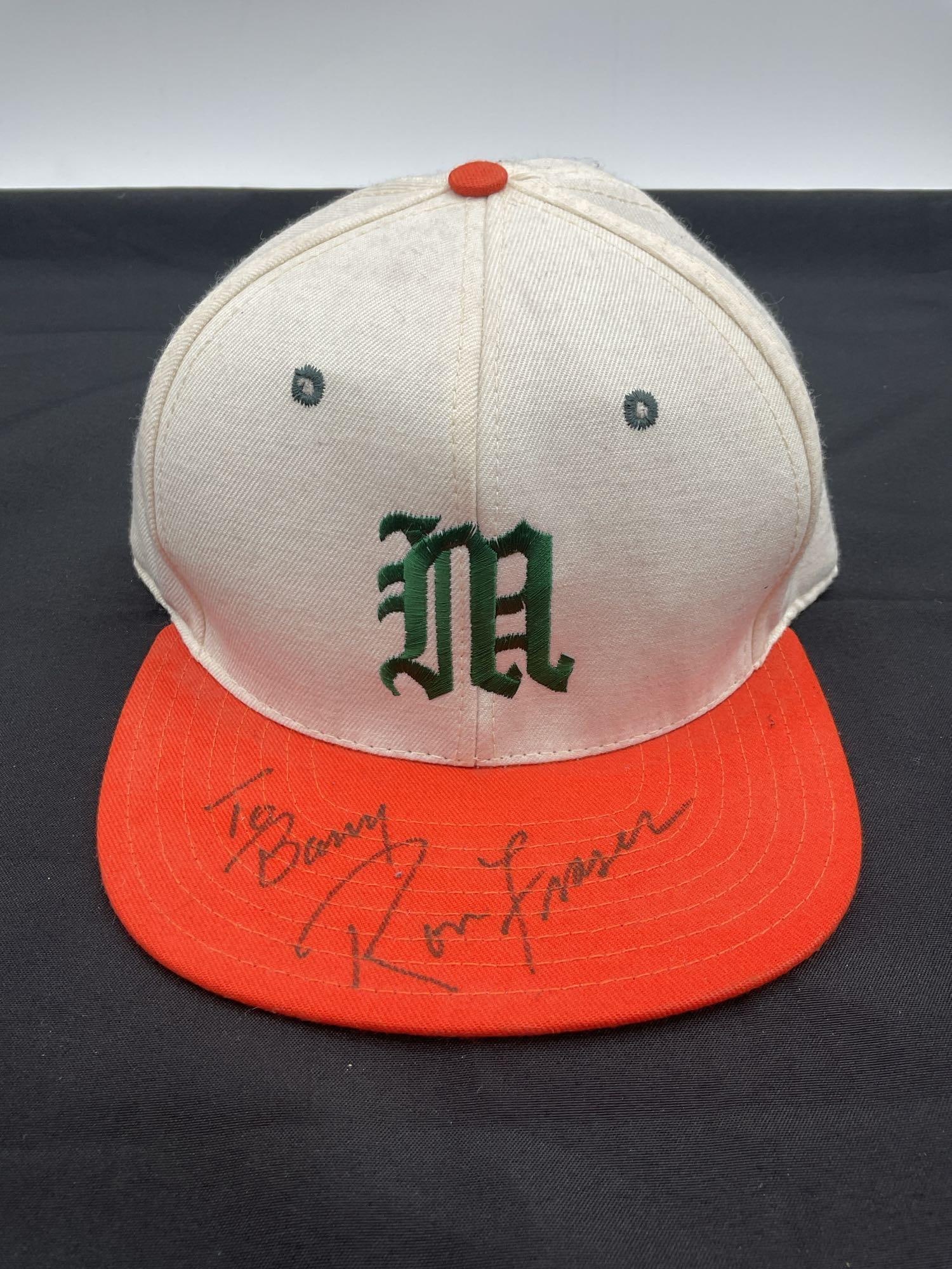 University of Miami Baseball Cap Signed by Ron Fraser: This vintage University of Miami baseball cap features the iconic green "M" logo on a white six-panel design with an orange brim. It is personally signed by legendary coach Ron Fraser with a