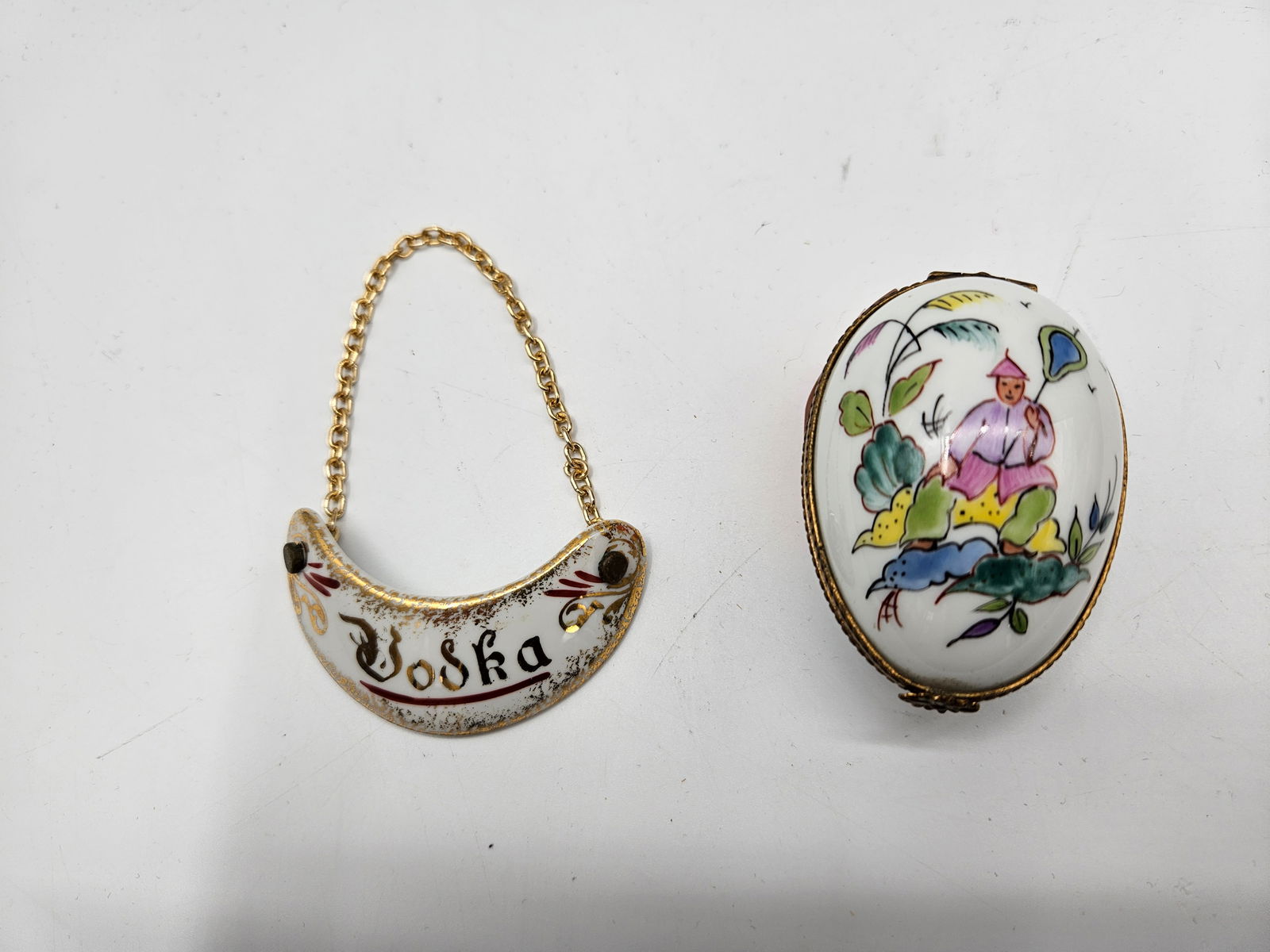 Limoges Hand-Painted Porcelain Trinket Box and “Vodka?? Decanter Tag: A beautiful matched pair of Limoges porcelain items, including a hand-painted trinket box made for Tiffany & Co. featuring a colorful pastoral scene, and a Limoges “Vodka” decanter tag