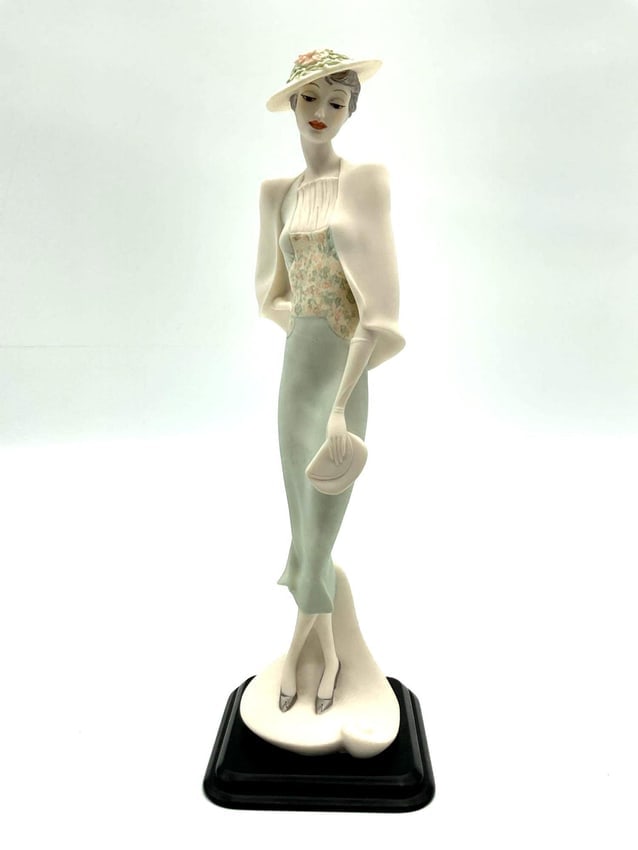 Art Deco Style Lady with Hat and Handbag Figurine: Graceful figurine of a poised woman in a pastel green and floral outfit, wearing a wide-brimmed hat adorned with flowers and holding a small handbag. Styled in the Art Deco tradition. Dimensions: H12i