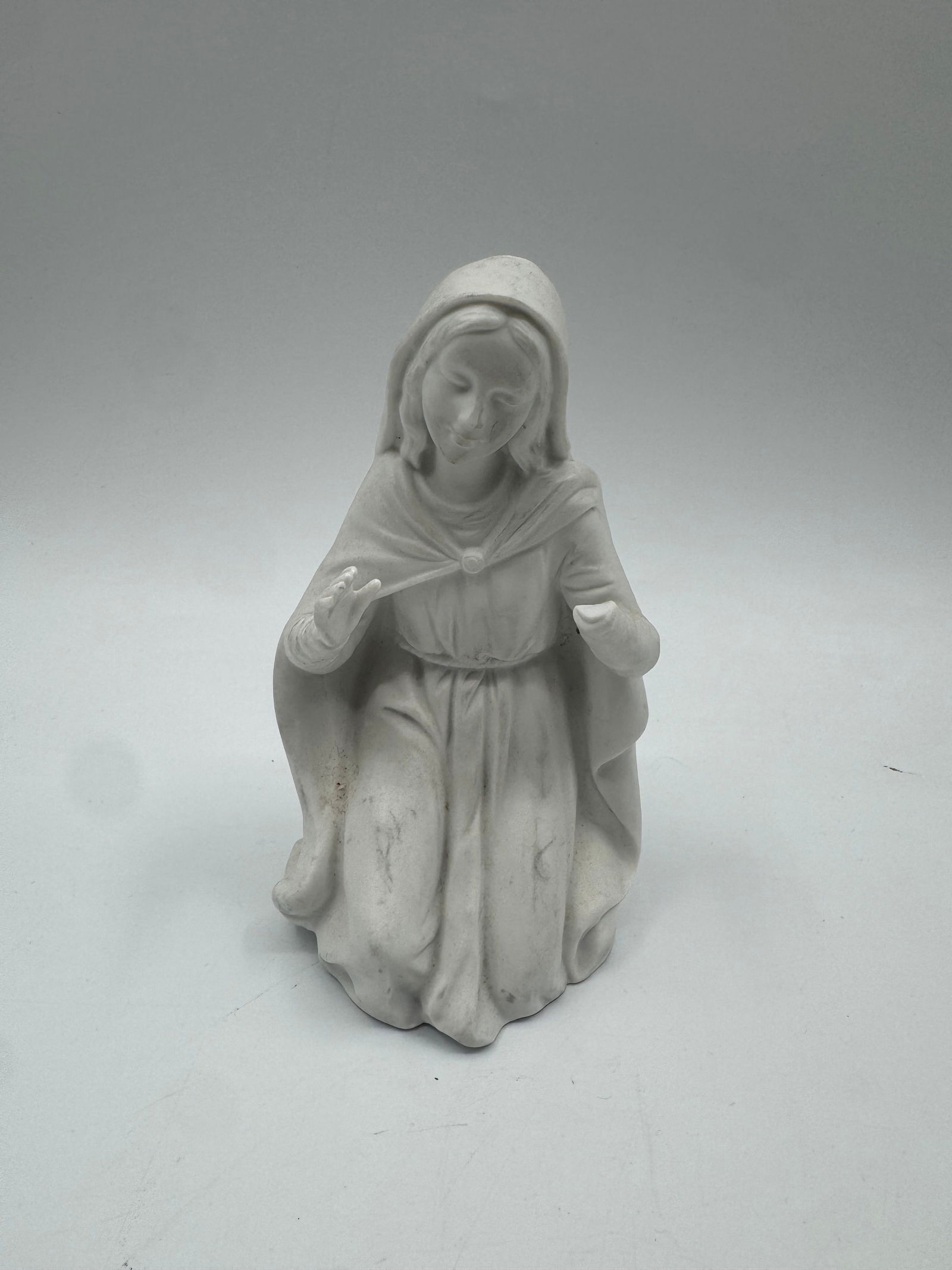 Bisque Porcelain Kneeling Madonna Figurine: Bisque porcelain figurine depicting the Virgin Mary kneeling in prayer. Marked on the base. Dimensions: 6" H x 3.5" L x 3.5" W.