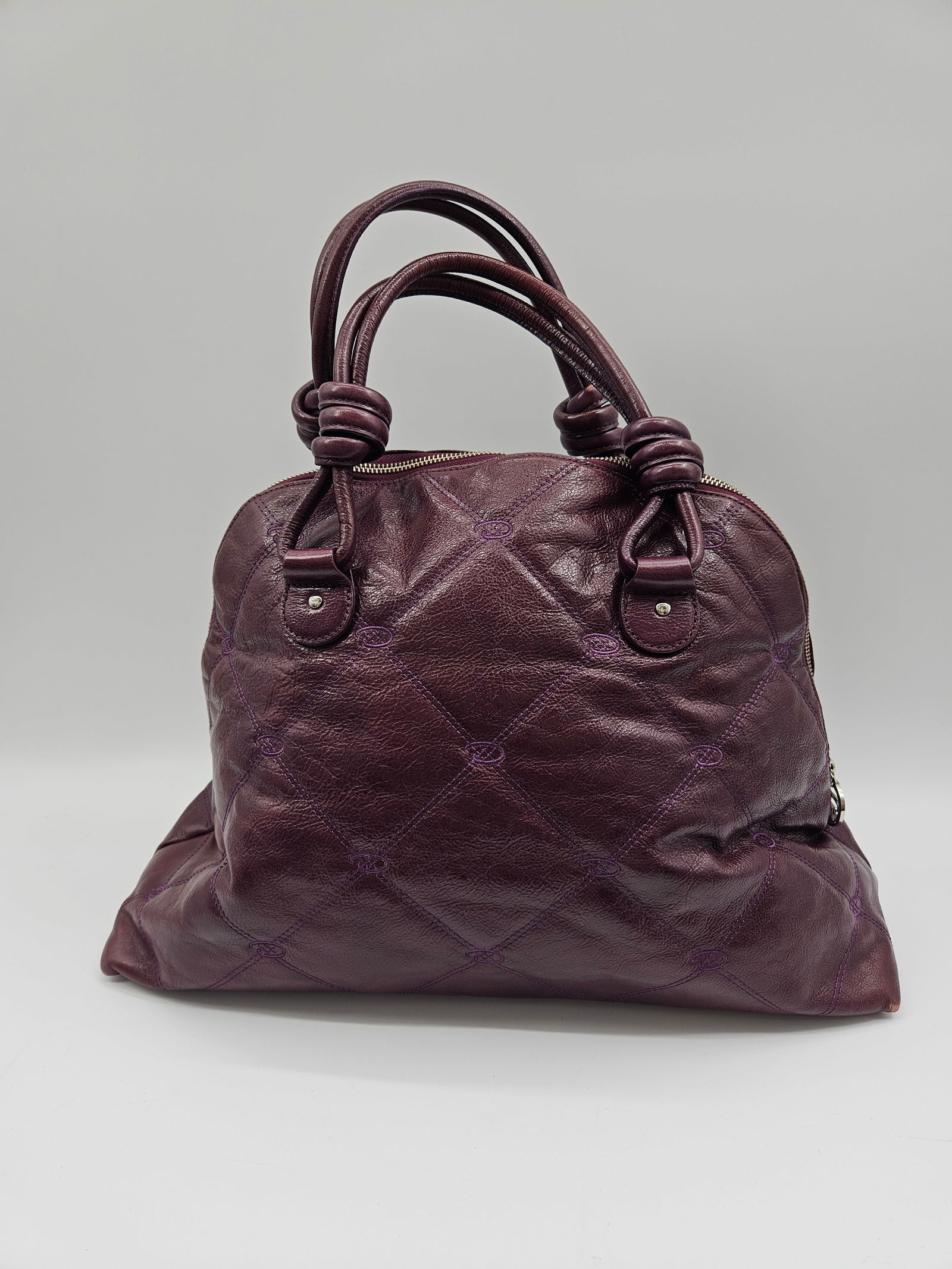 Oscar de la Renta Quilted Leather Handbag: Oscar de la Renta quilted leather handbag in a deep purple hue. Features double handles with knotted details and a red fabric interior. Dimensions: 17 in height with handles, 17.5 in width, 12 in heig
