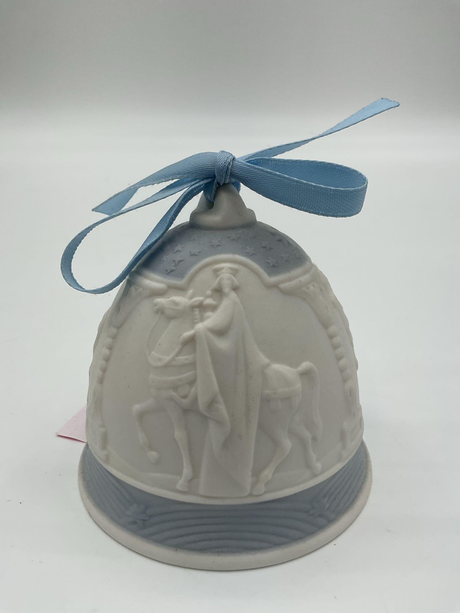 Lladro 1990 Christmas Bell Ornament: A matte porcelain bell featuring a Three Kings motif in bas-relief, with one king standing beside a horse, topped with a blue ribbon, and dated 1990. Dimensions: H3in Dia3in Medium: Unglazed (matte) p